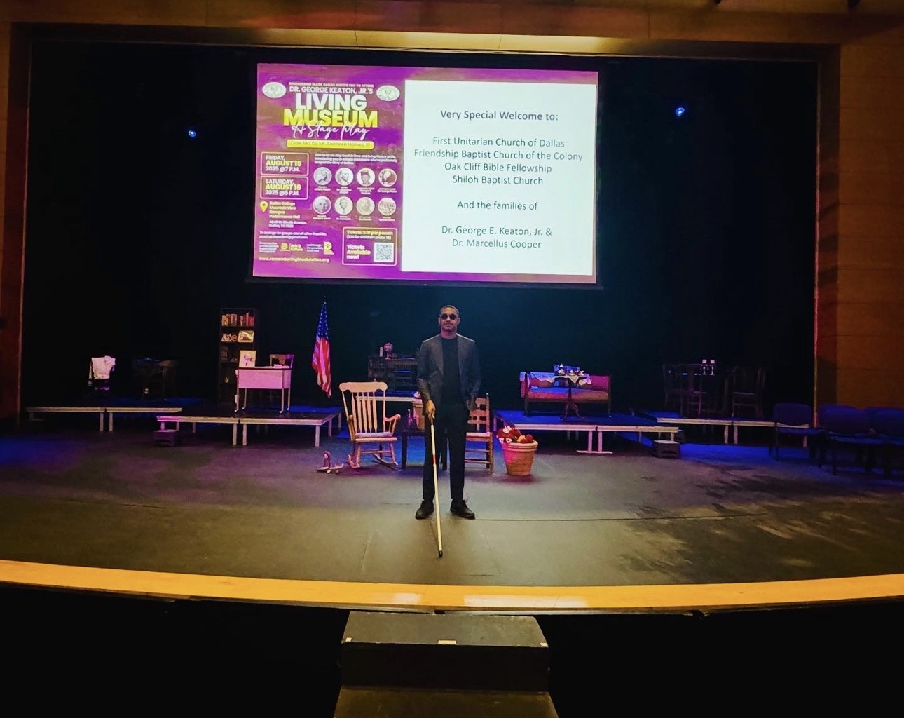 Person standing on stage in front of a large screen with presentation slide, stage set with chairs and American flag, auditorium setting.