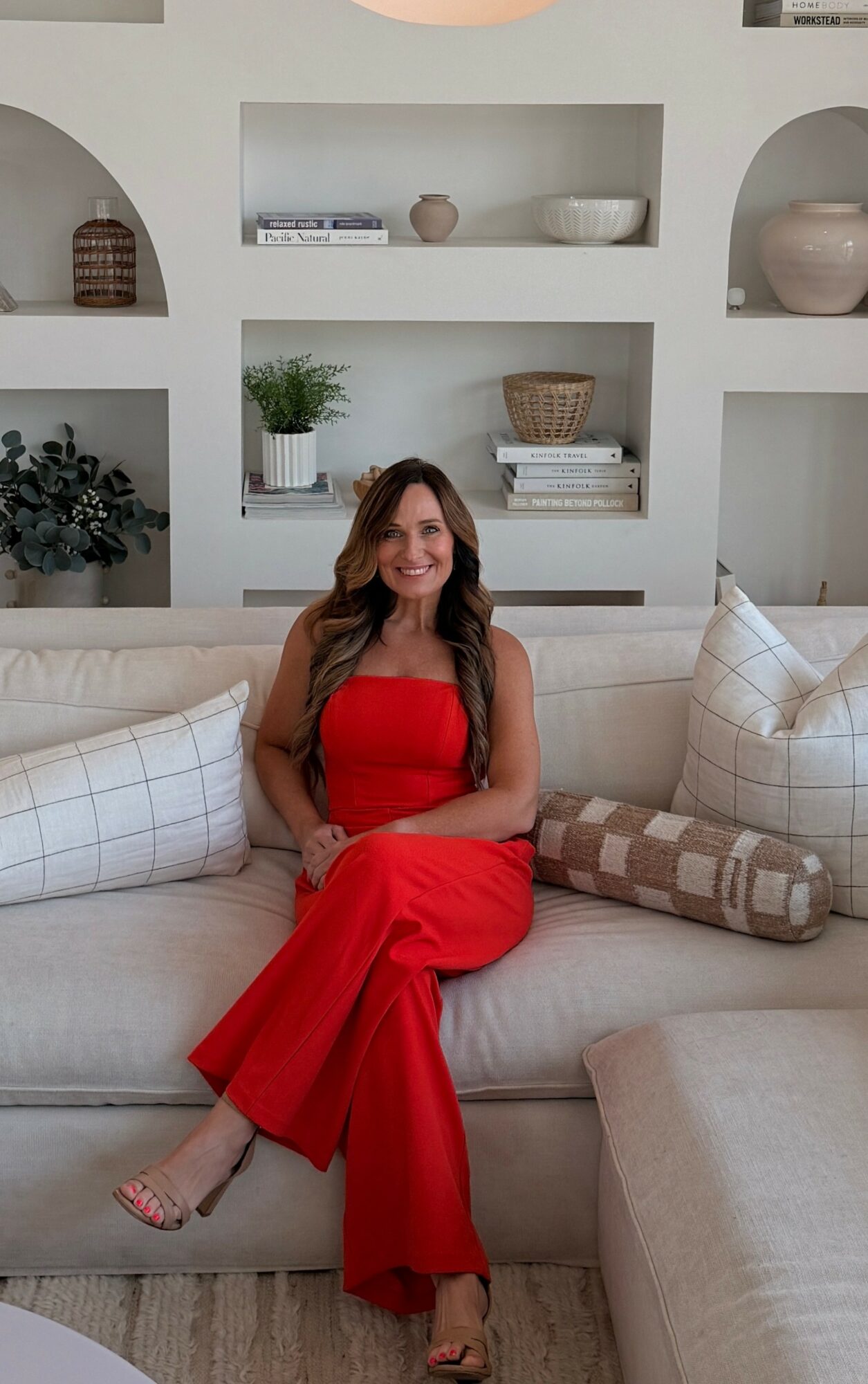 Woman in red jumpsuit sitting on a beige sofa in a room with white shelves and decorative items behind her.