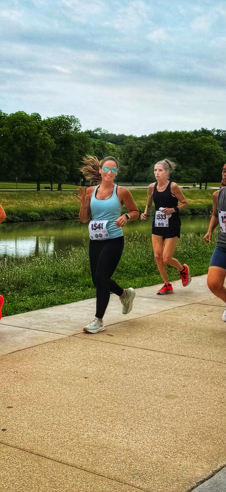Group of people running outdoors on a paved path near a pond with trees and cloudy sky in background.