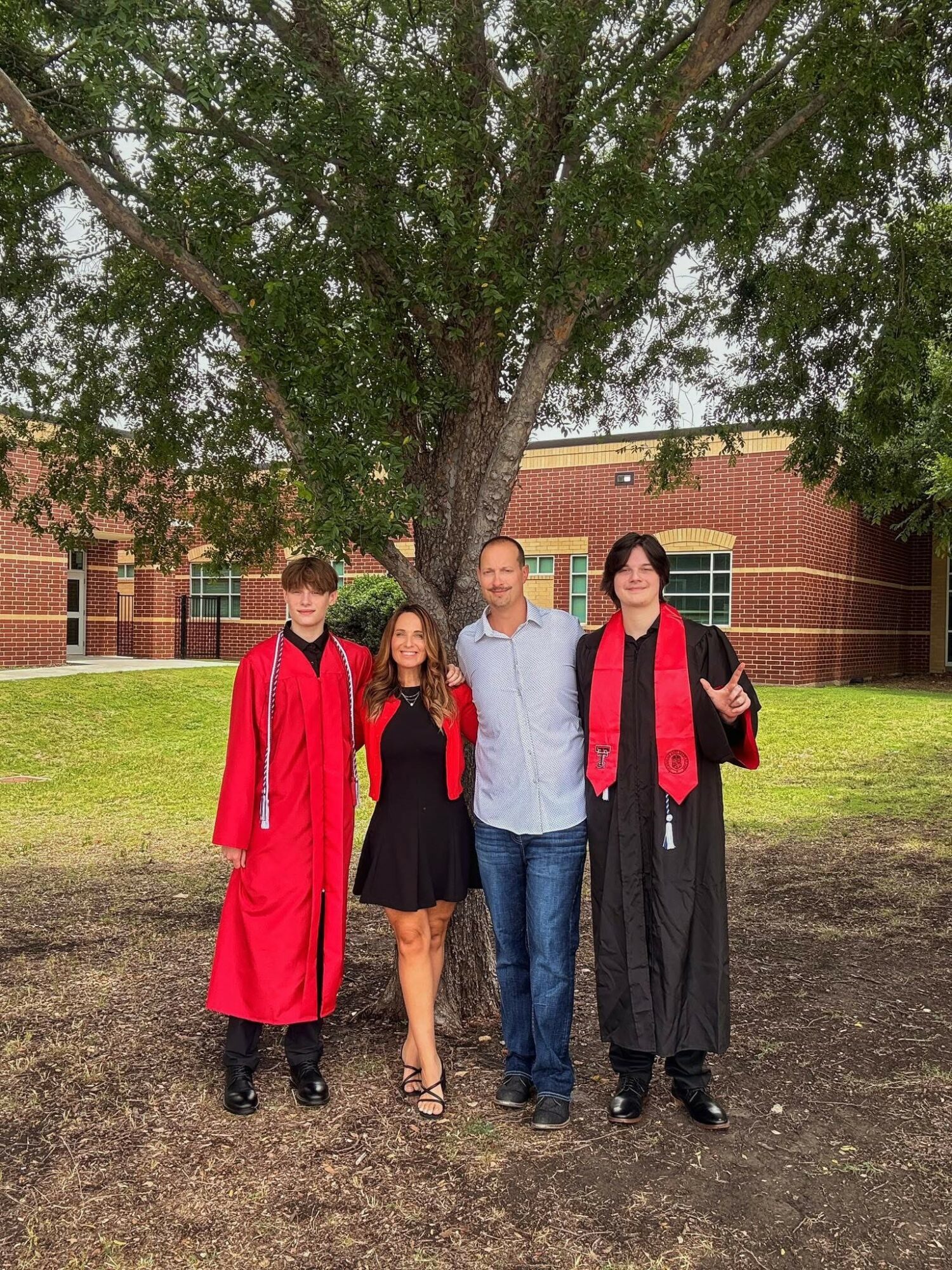 Four people standing outdoors in front of a large tree and brick building, smiling, with grass and dirt ground.