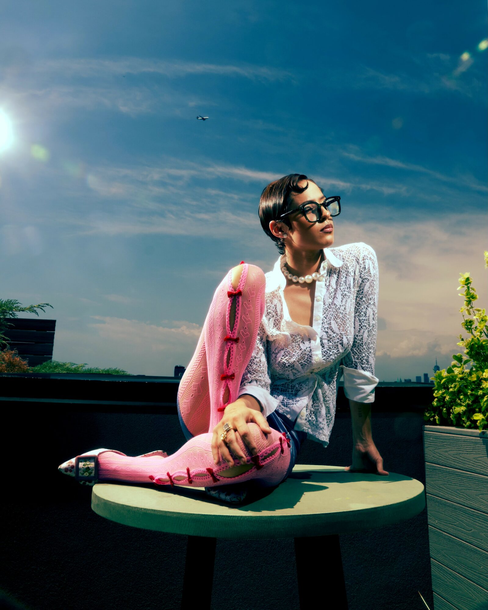 Woman with short hair, glasses, and pearl necklace sitting outdoors at a table with a pink stuffed animal, under a blue sky.