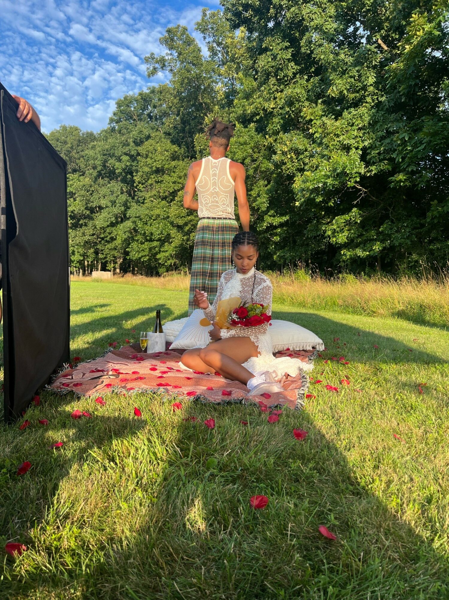 Child sitting on a blanket with flowers, with a woman standing behind in a grassy park with trees and blue sky.