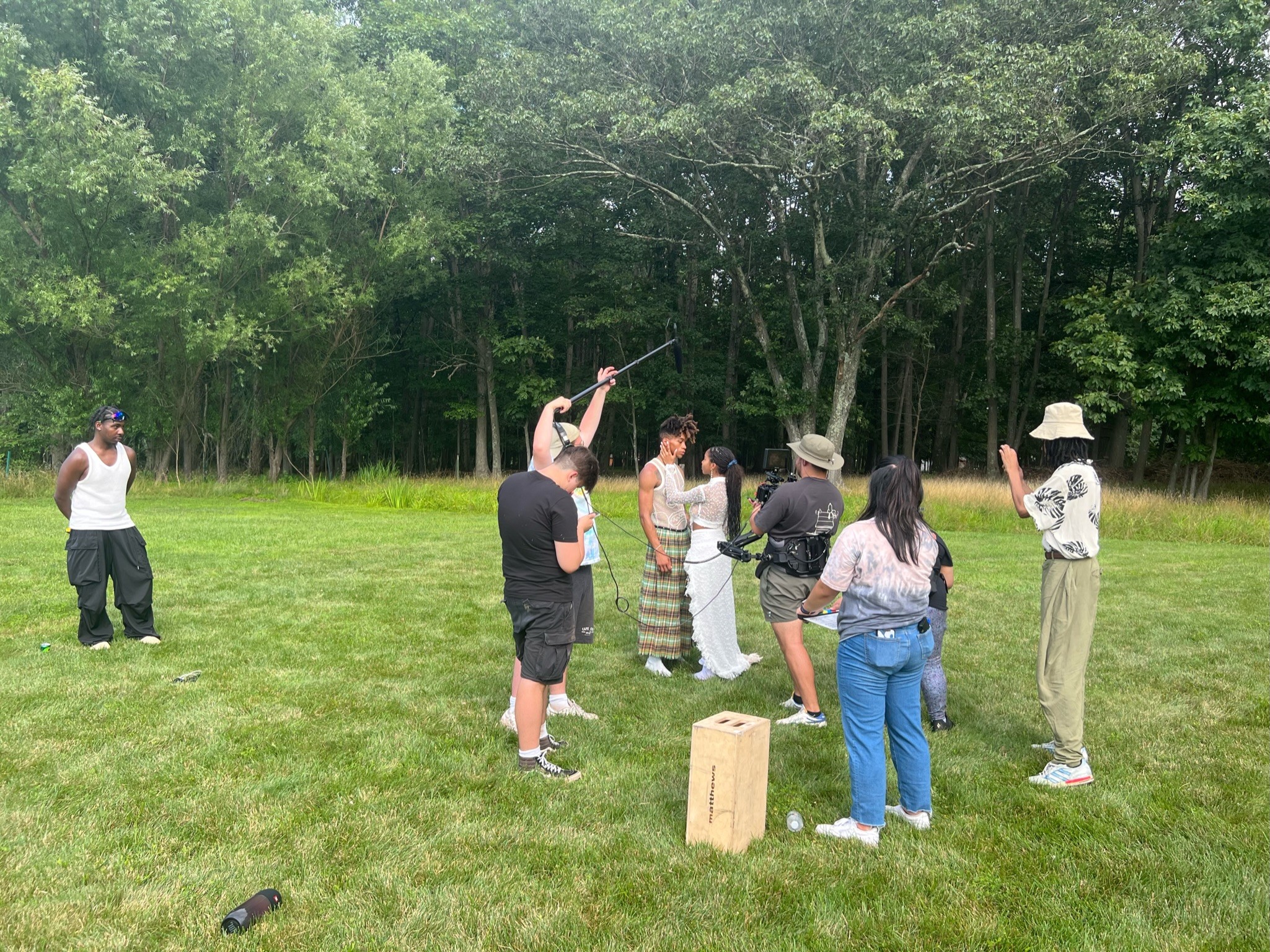 Group of people outdoors on grass, some watching and others participating in an activity, with trees in the background.