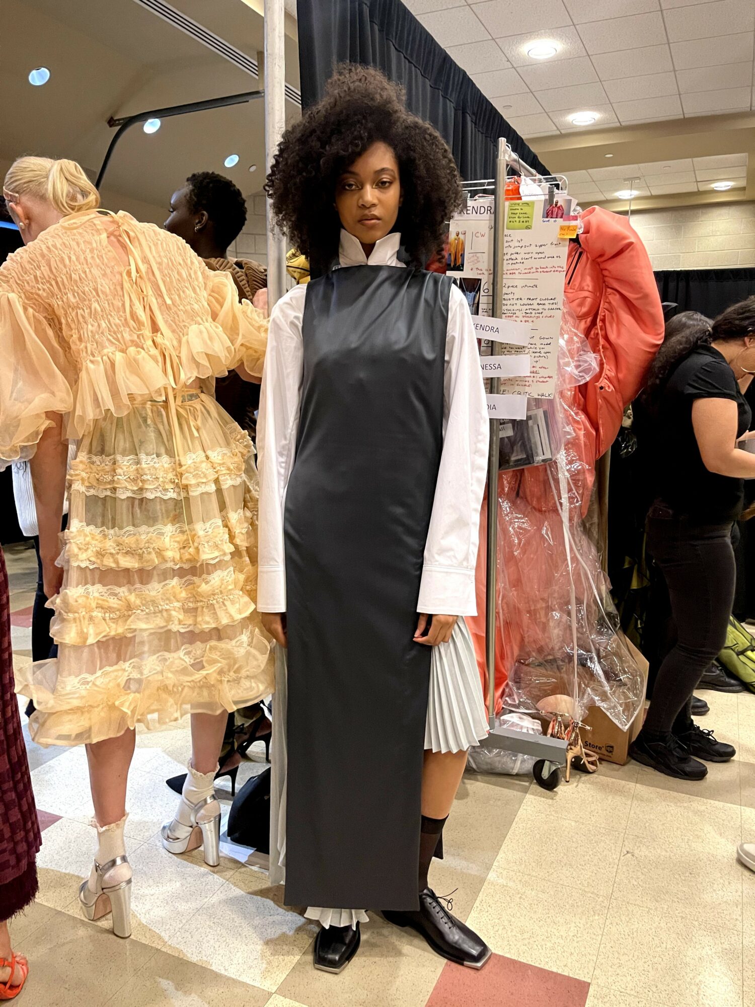 Young woman with curly hair in a black dress and white shirt, standing in a busy indoor setting.