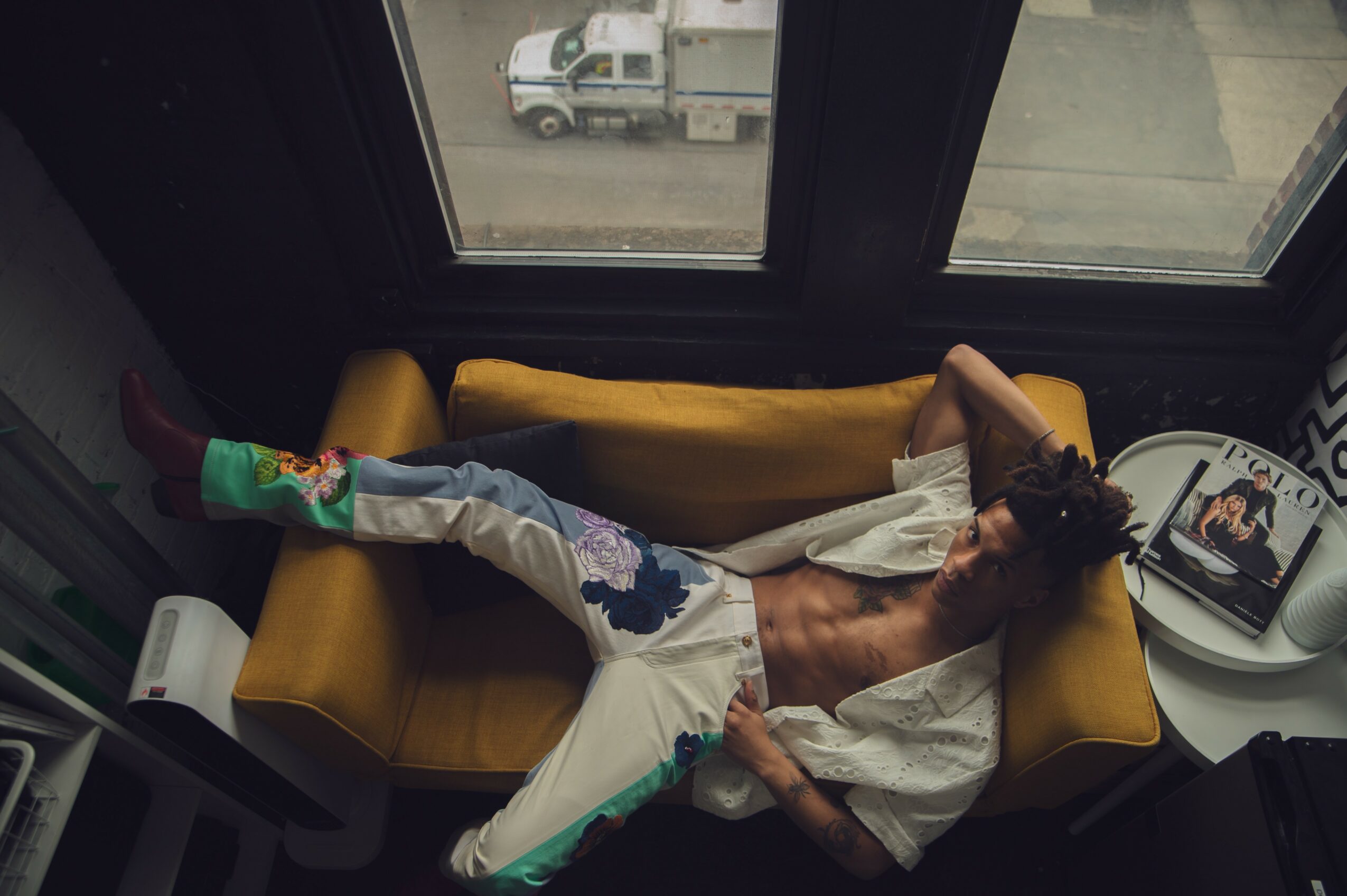 Person lying on a yellow couch near a window, wearing colorful socks and white pants, with a magazine nearby.