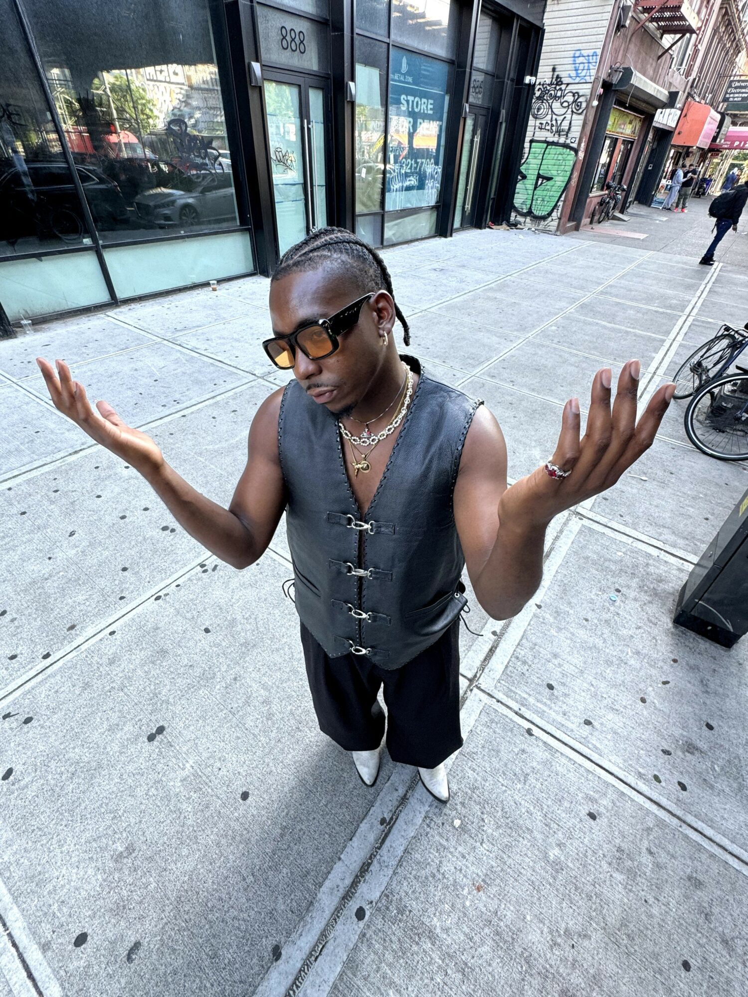 Person with short hair, sunglasses, and jewelry stands on sidewalk with arms raised, buildings and bicycles in background.