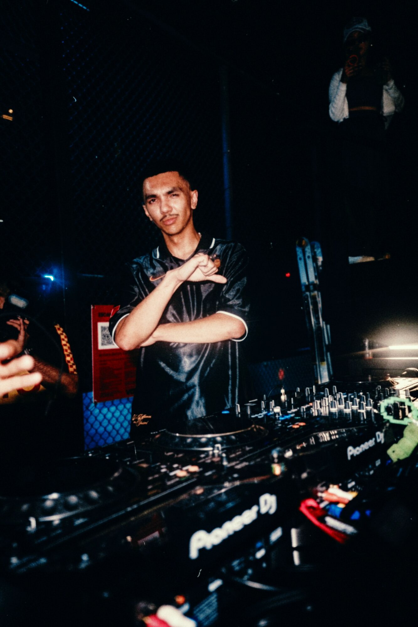 Young man DJing at a club with equipment, dark background, and a person standing in the background, smiling confidently.
