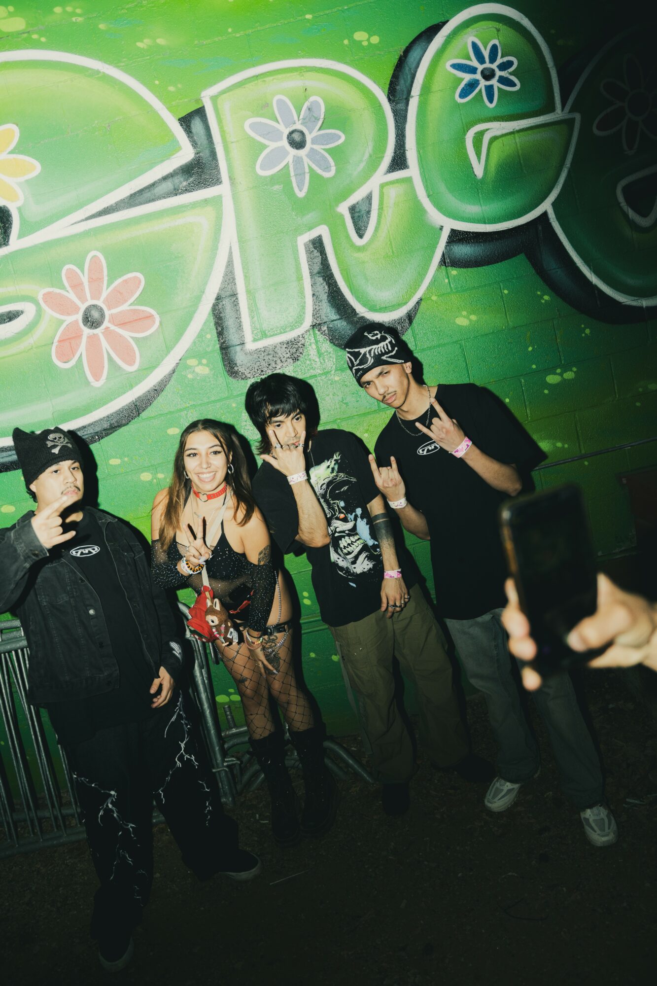 Four young people pose in front of a green graffiti wall with flowers, some making peace signs, in a dark setting.