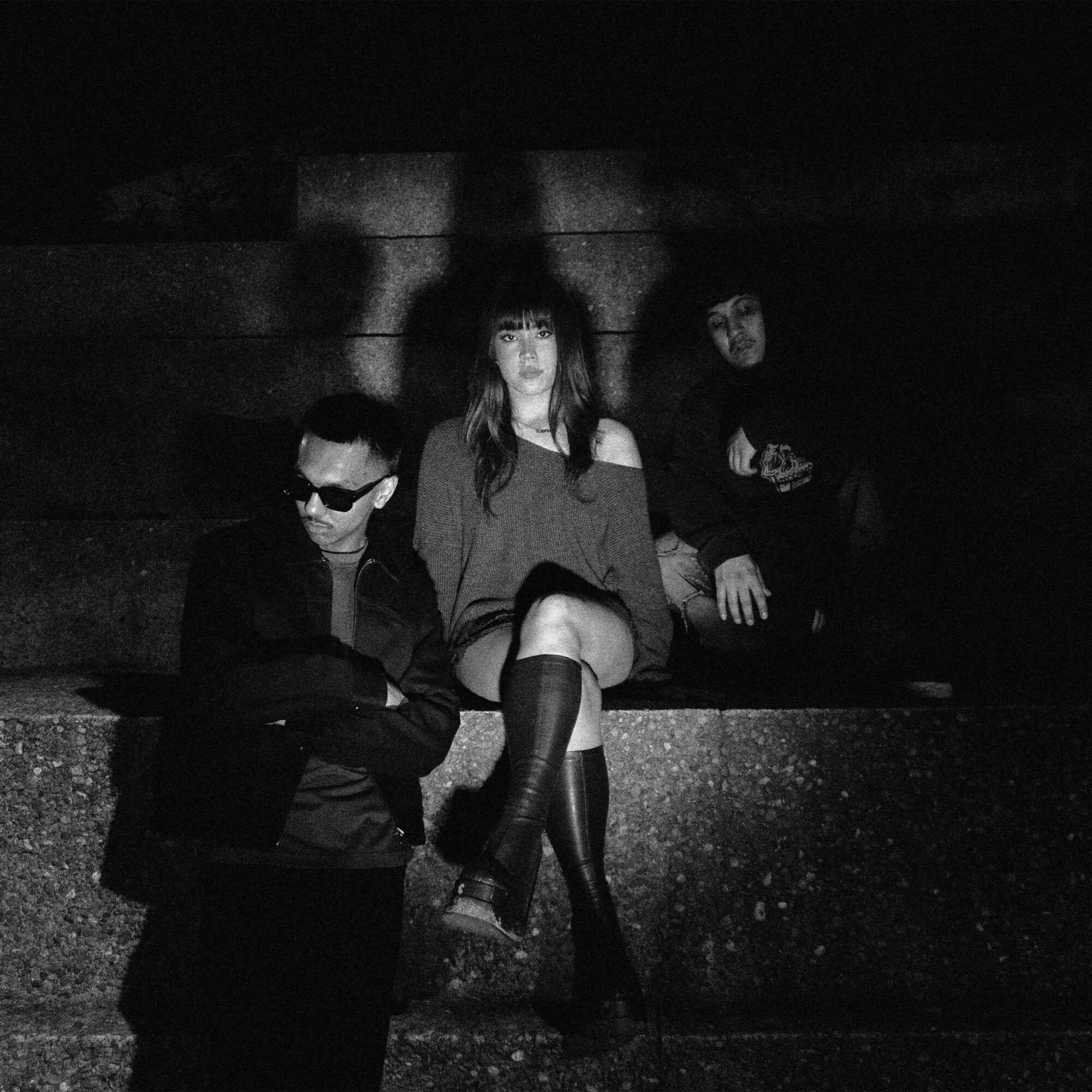 Three people sitting on steps in a dark setting, one woman in the center with crossed legs, two men on either side, all in dark clothing.