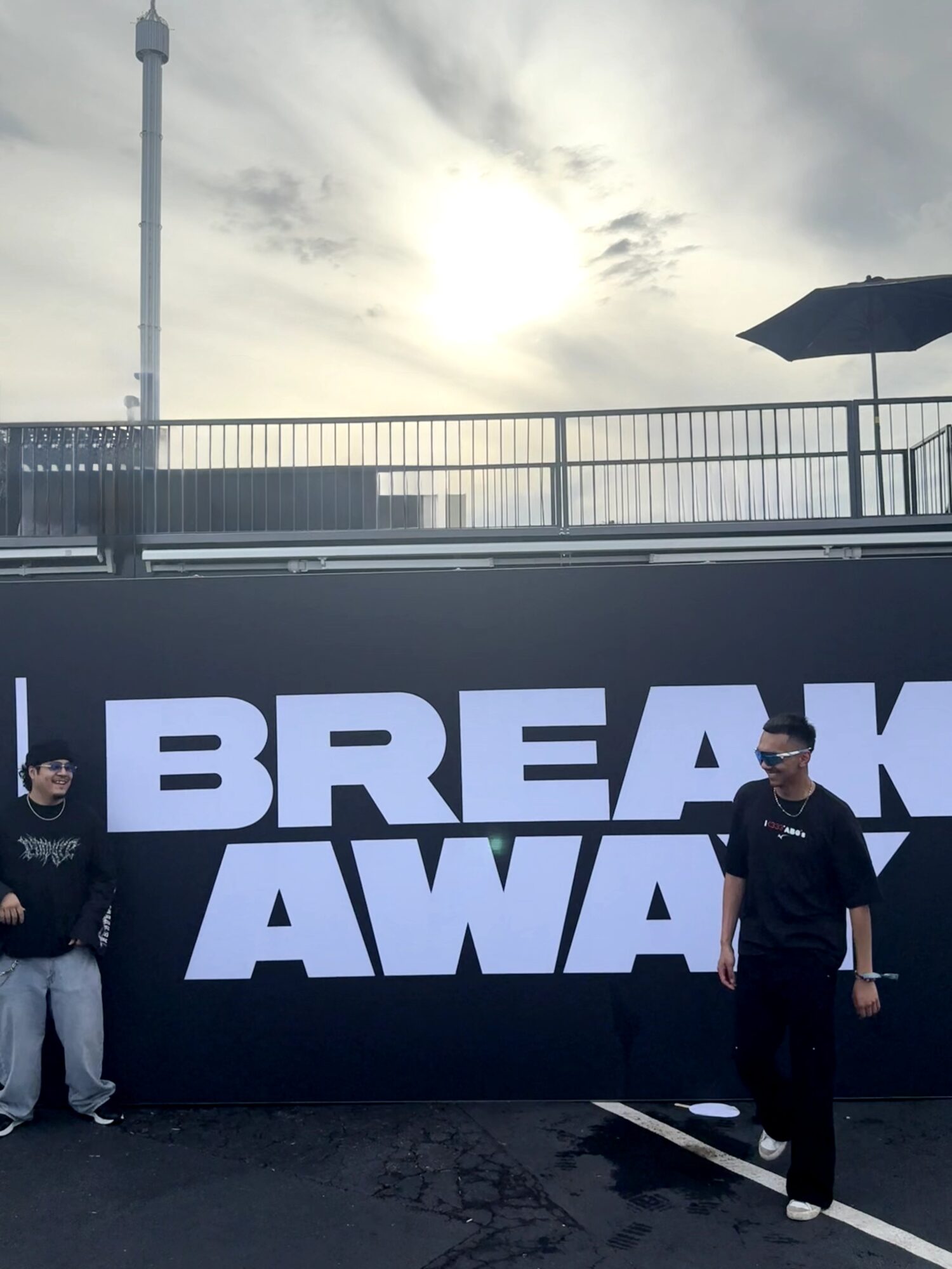 Two people standing in front of a large sign with the words 'BREAK AWAY' outdoors, with a cloudy sky and a railing in the background.