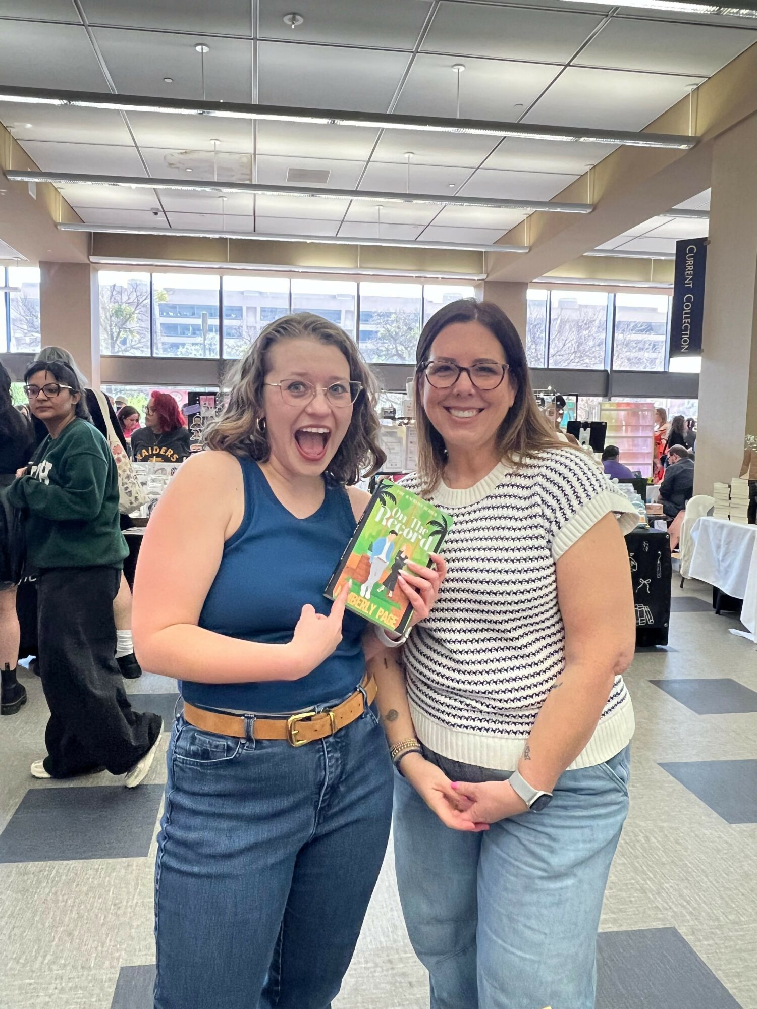 Two women smiling, one holding a colorful book, standing indoors with large windows and a checkered floor.