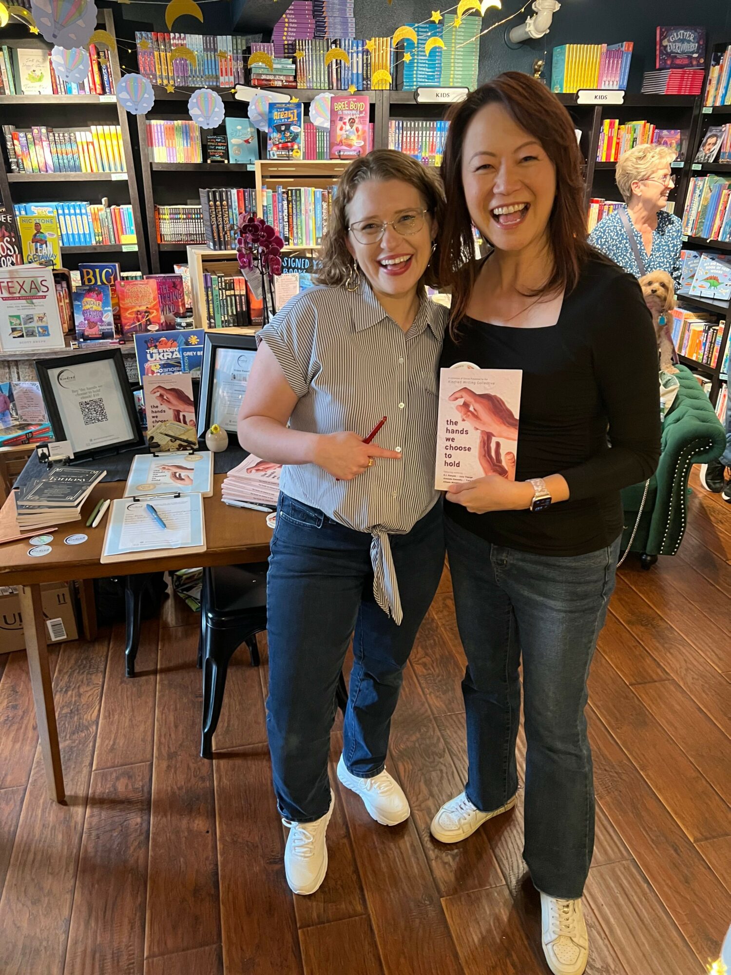 Two women standing in a bookstore, one holding a book, smiling, with shelves of books behind them.