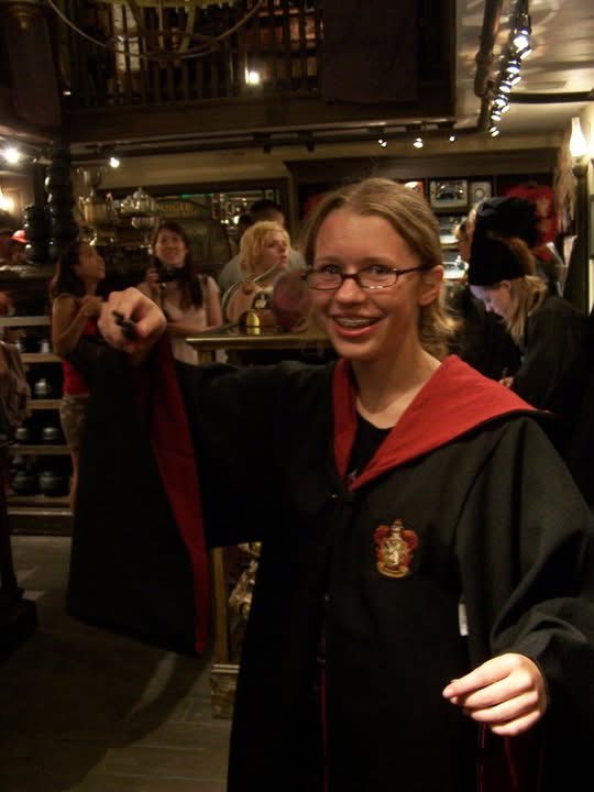 Person in a black gown with red accents and glasses smiling indoors, surrounded by people and warm lighting.