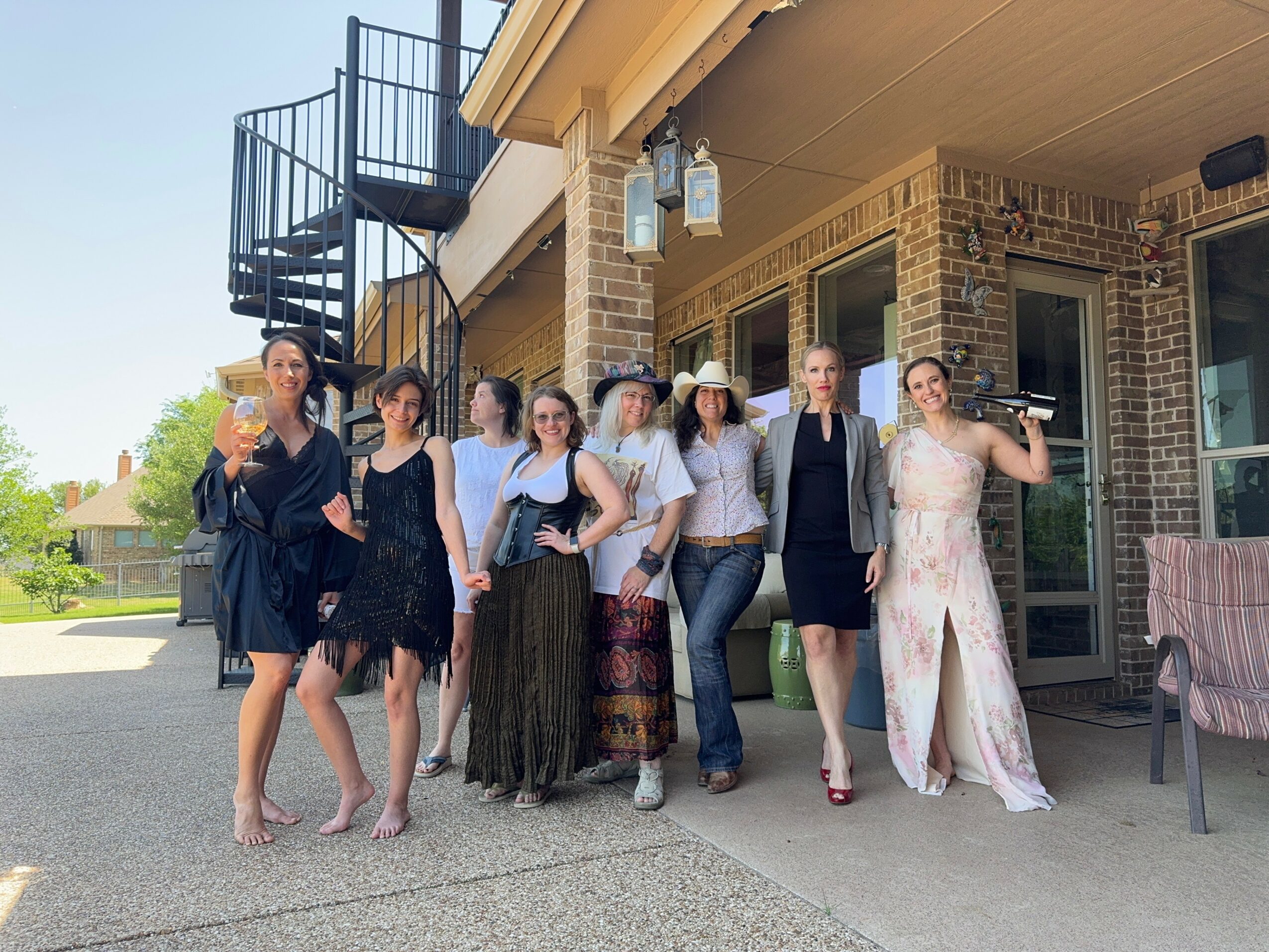 Seven women stand outside a building, some holding drinks, dressed in formal and casual attire, smiling and posing.