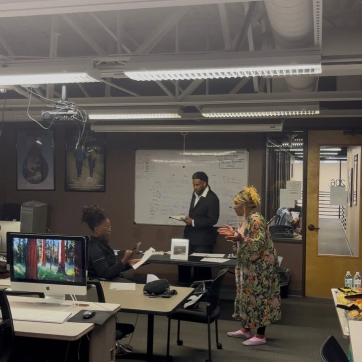 Three people in a classroom or meeting room, one standing and two seated, with a whiteboard and computer screens visible.