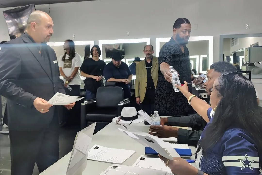 People in a room, some seated at a table, others standing, with a person handing a document to a seated woman.