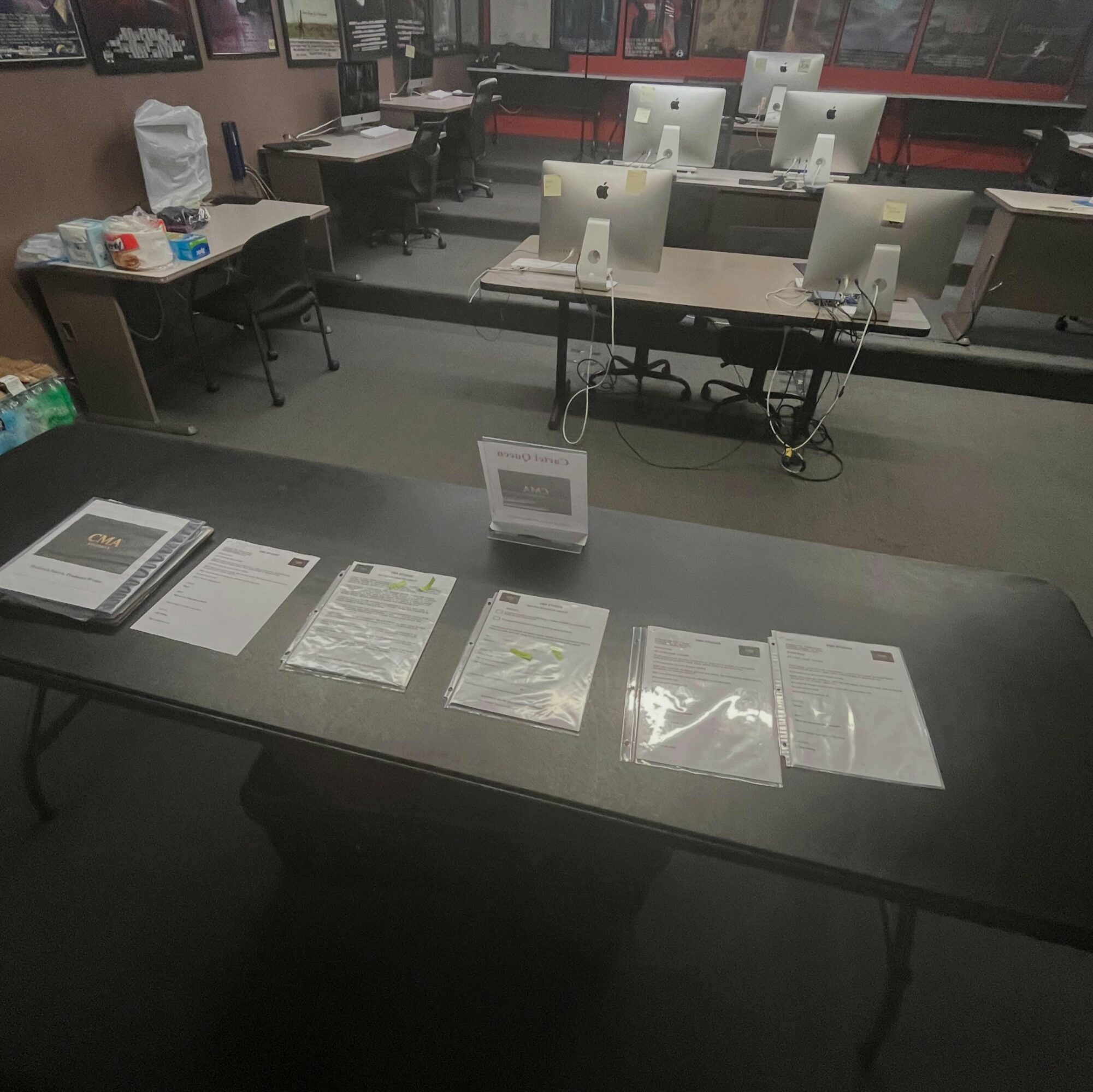 Classroom with desks, chairs, and computers, some papers and a sign on a table in the foreground.