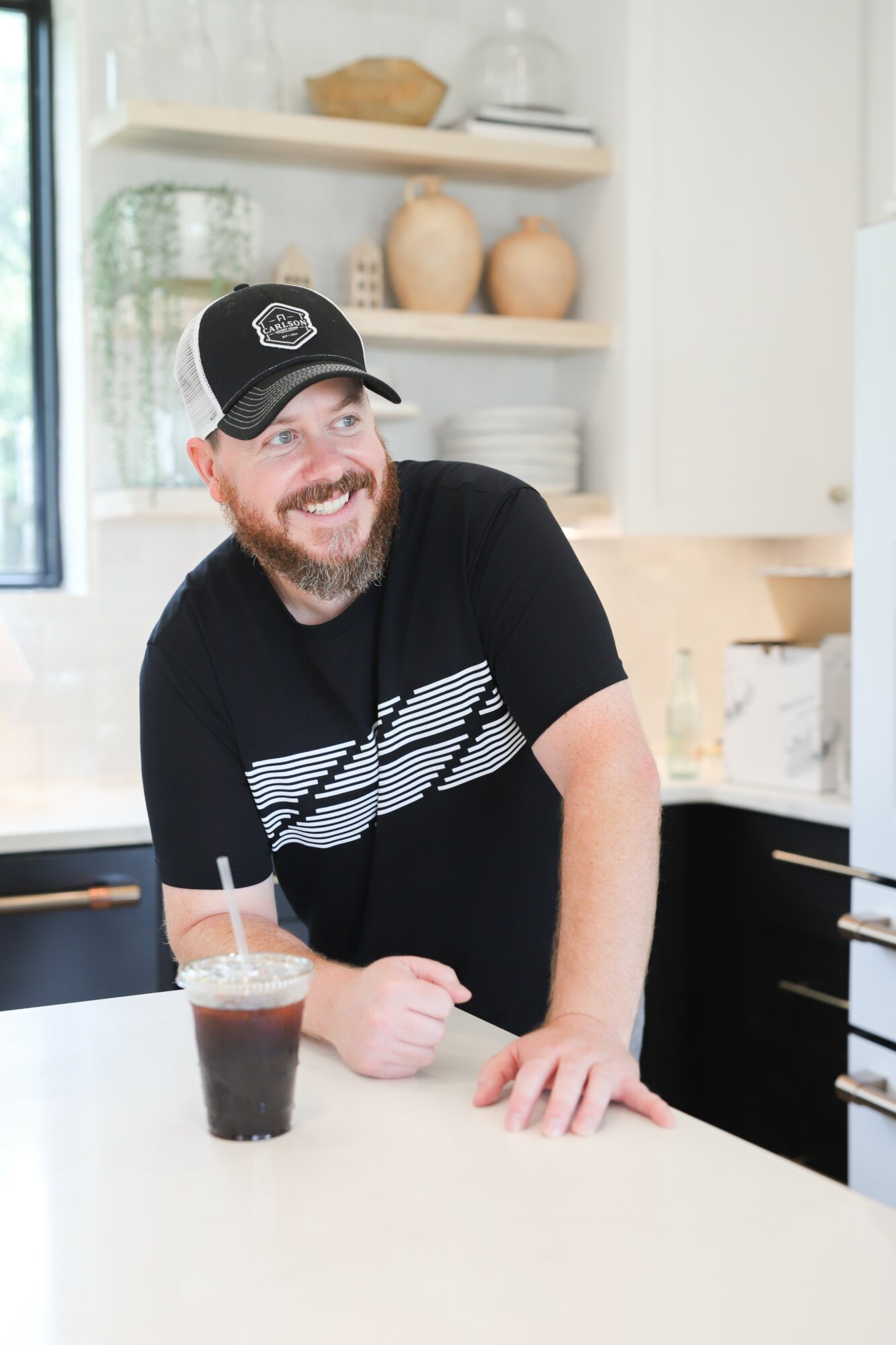 Smiling man with a beard wearing a black cap and T-shirt, leaning on a kitchen counter with a drink, in a bright kitchen.