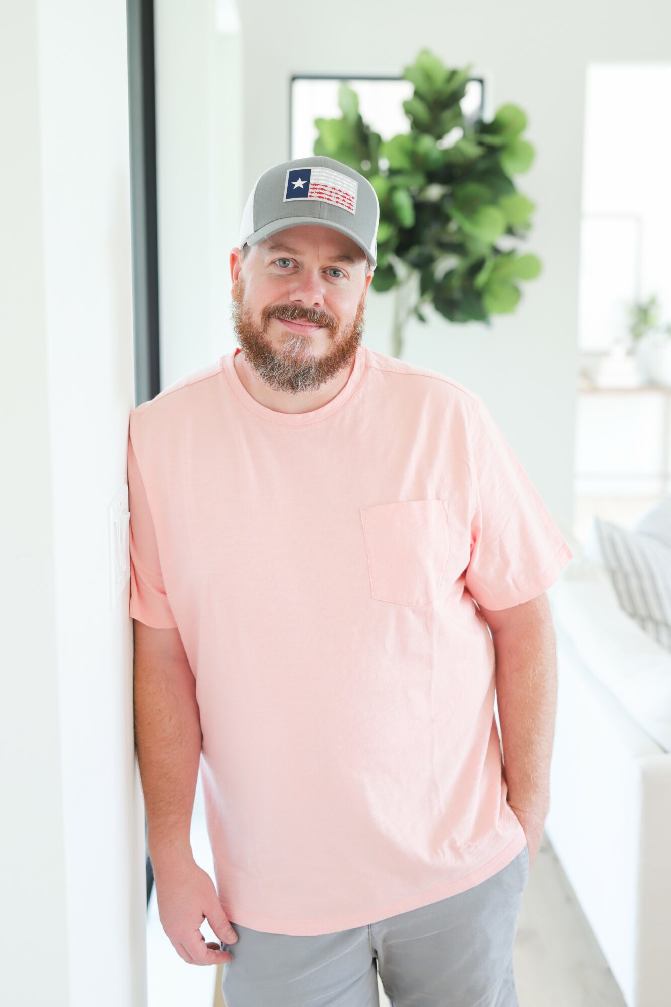 Man with a beard wearing a gray cap and pink T-shirt, standing indoors near a window with a plant in the background.