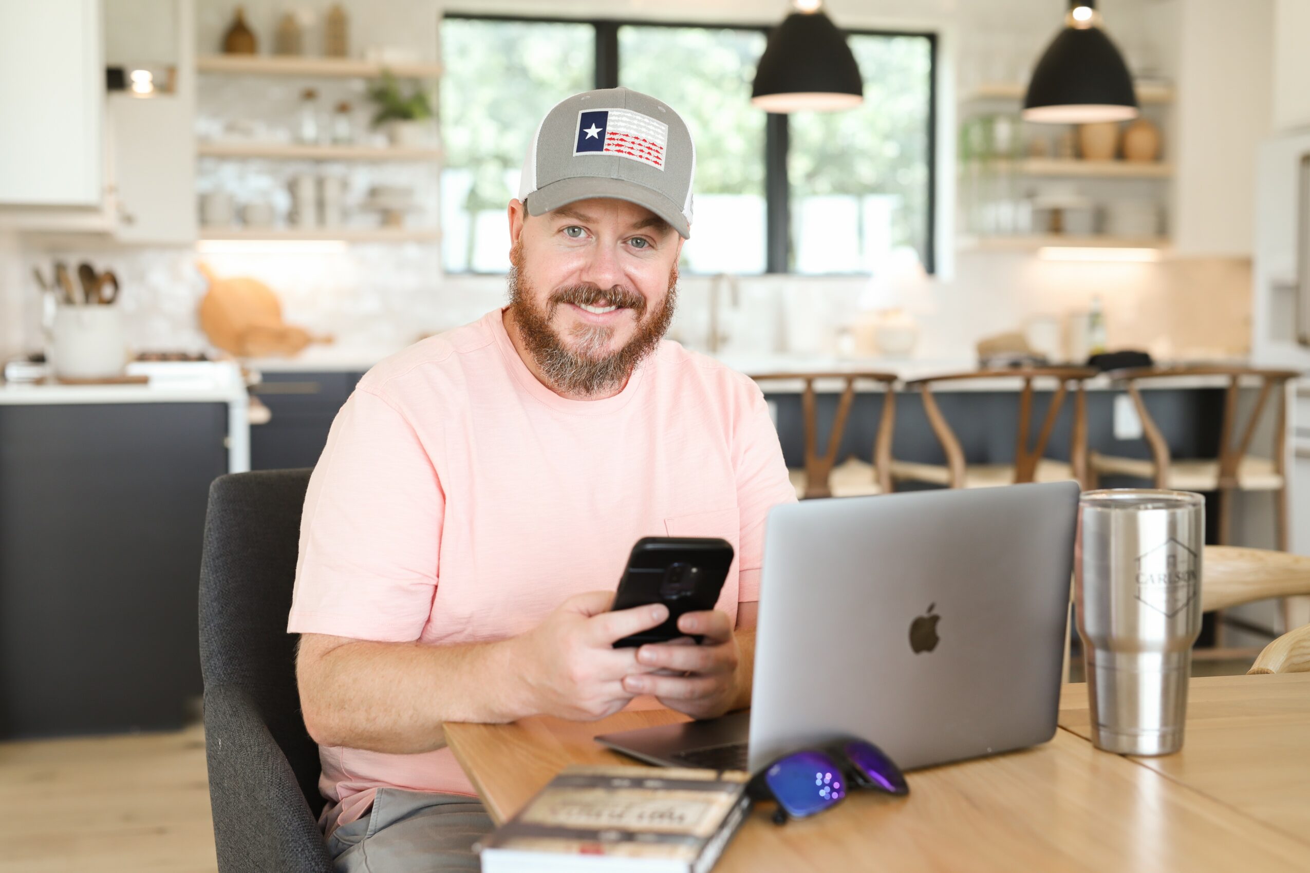 Man with a beard wearing a cap, sitting at a table with a laptop, holding a phone, in a bright kitchen.