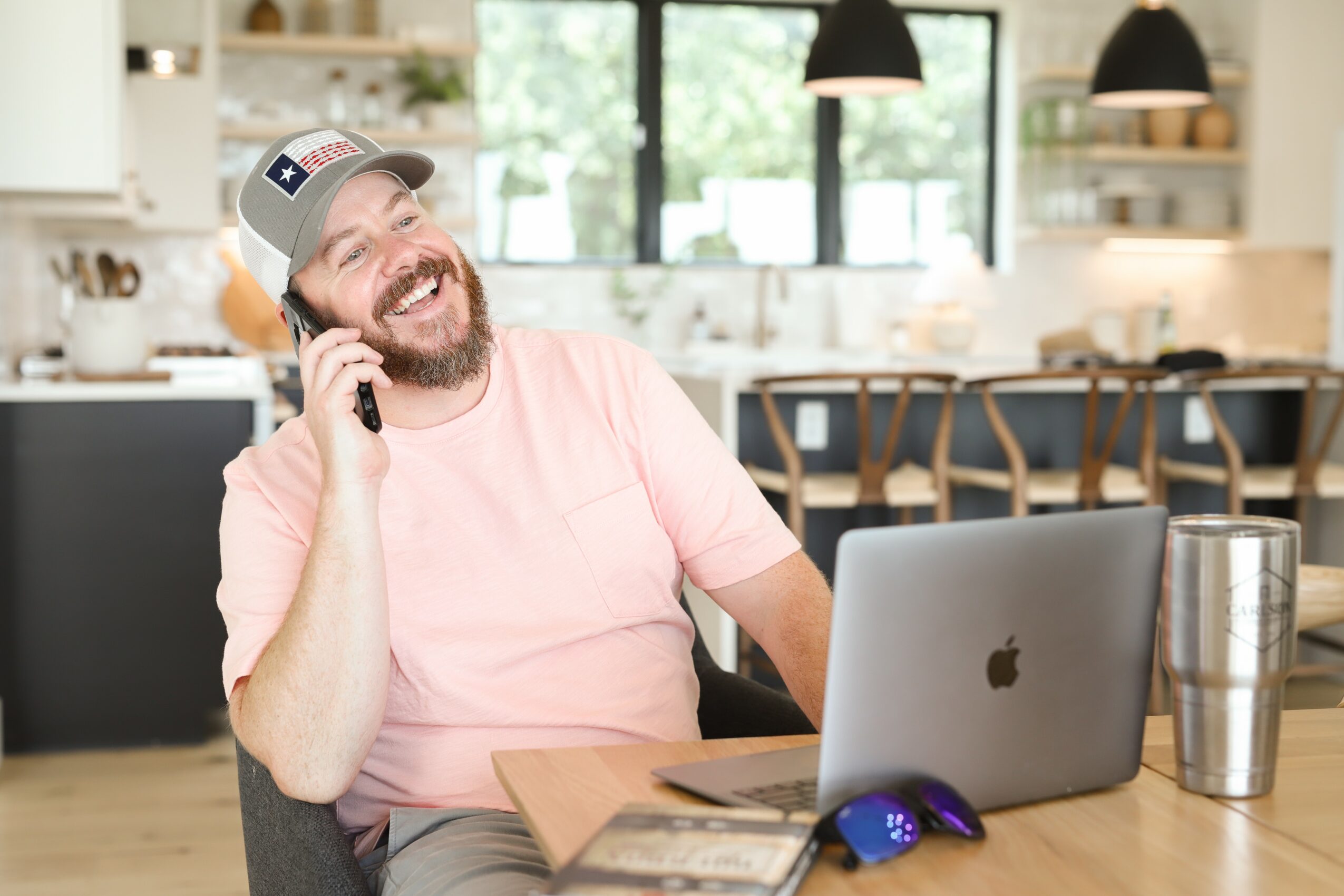 Man with a beard, wearing a cap, smiling, talking on a phone, sitting at a desk with a laptop, in a bright kitchen.