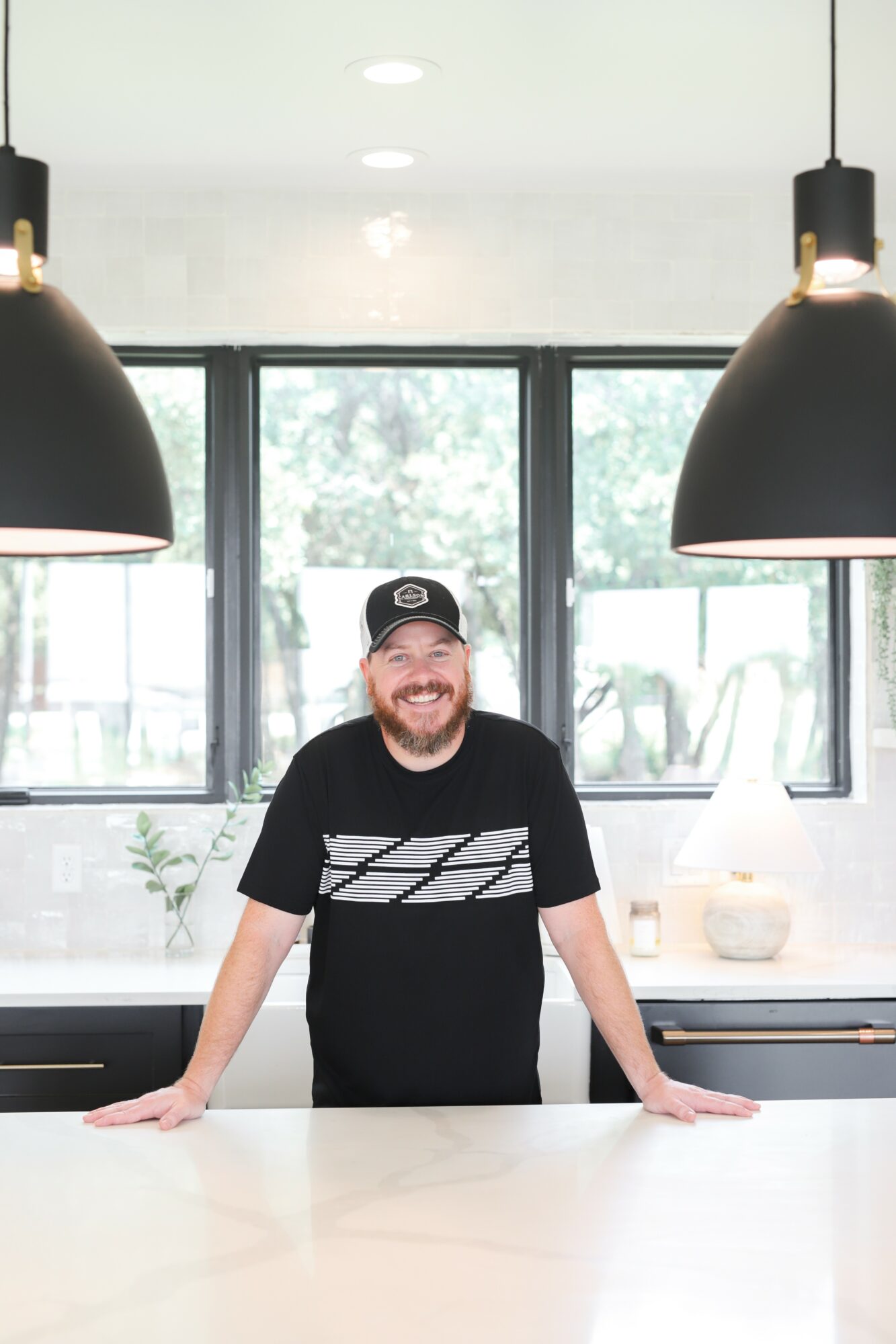 Man with a beard wearing a black cap and T-shirt, smiling, standing behind a white counter in a modern kitchen.