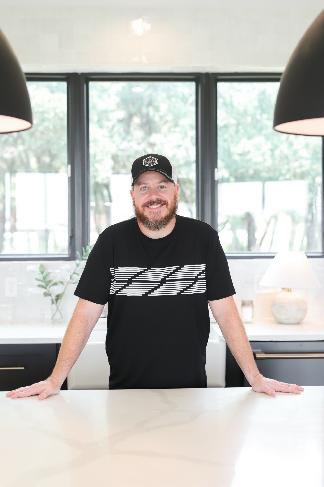 Man with a beard wearing a black cap and T-shirt, standing behind a white counter in a bright room.