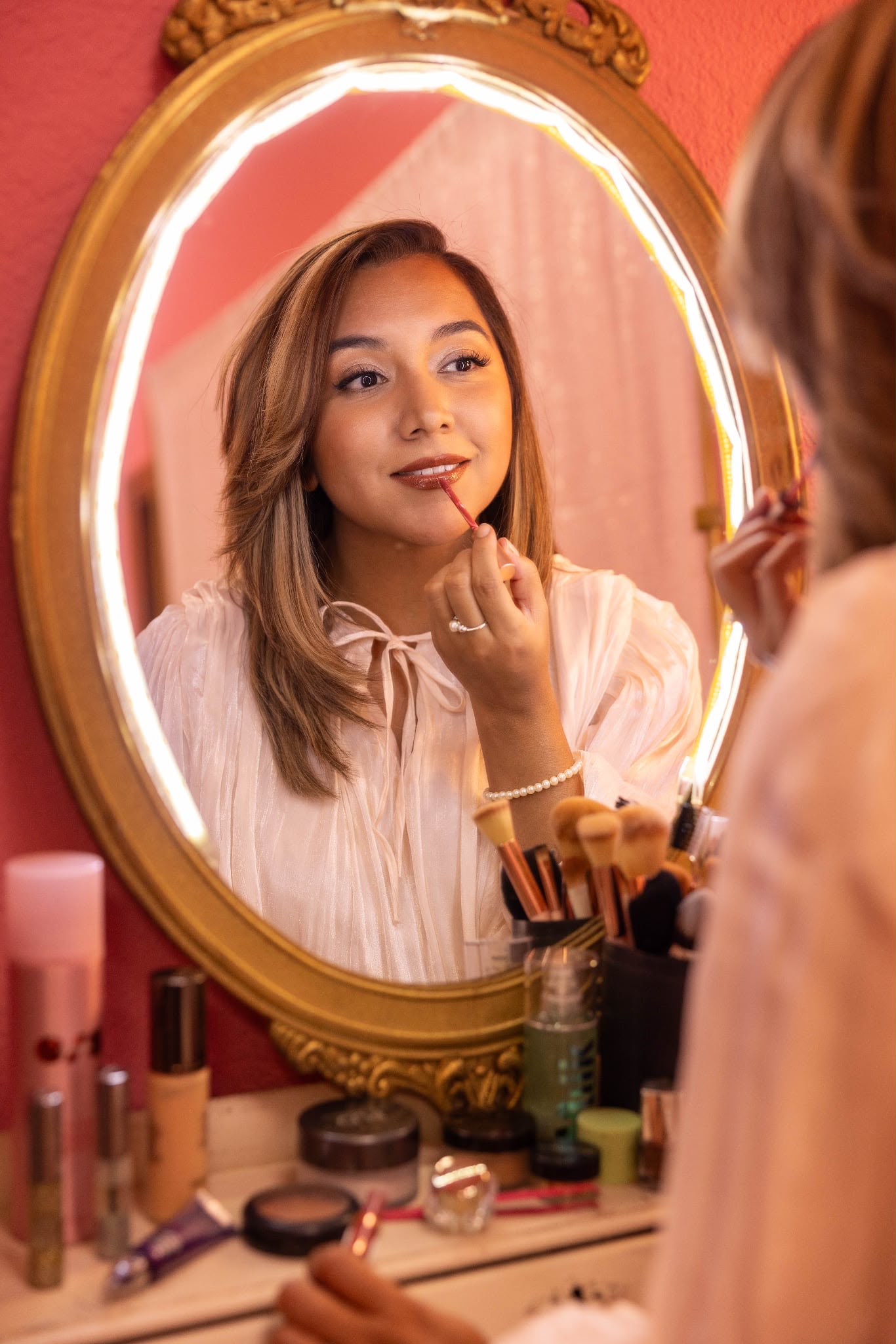 Young woman looking at her reflection in an oval mirror, touching her face, surrounded by makeup products.