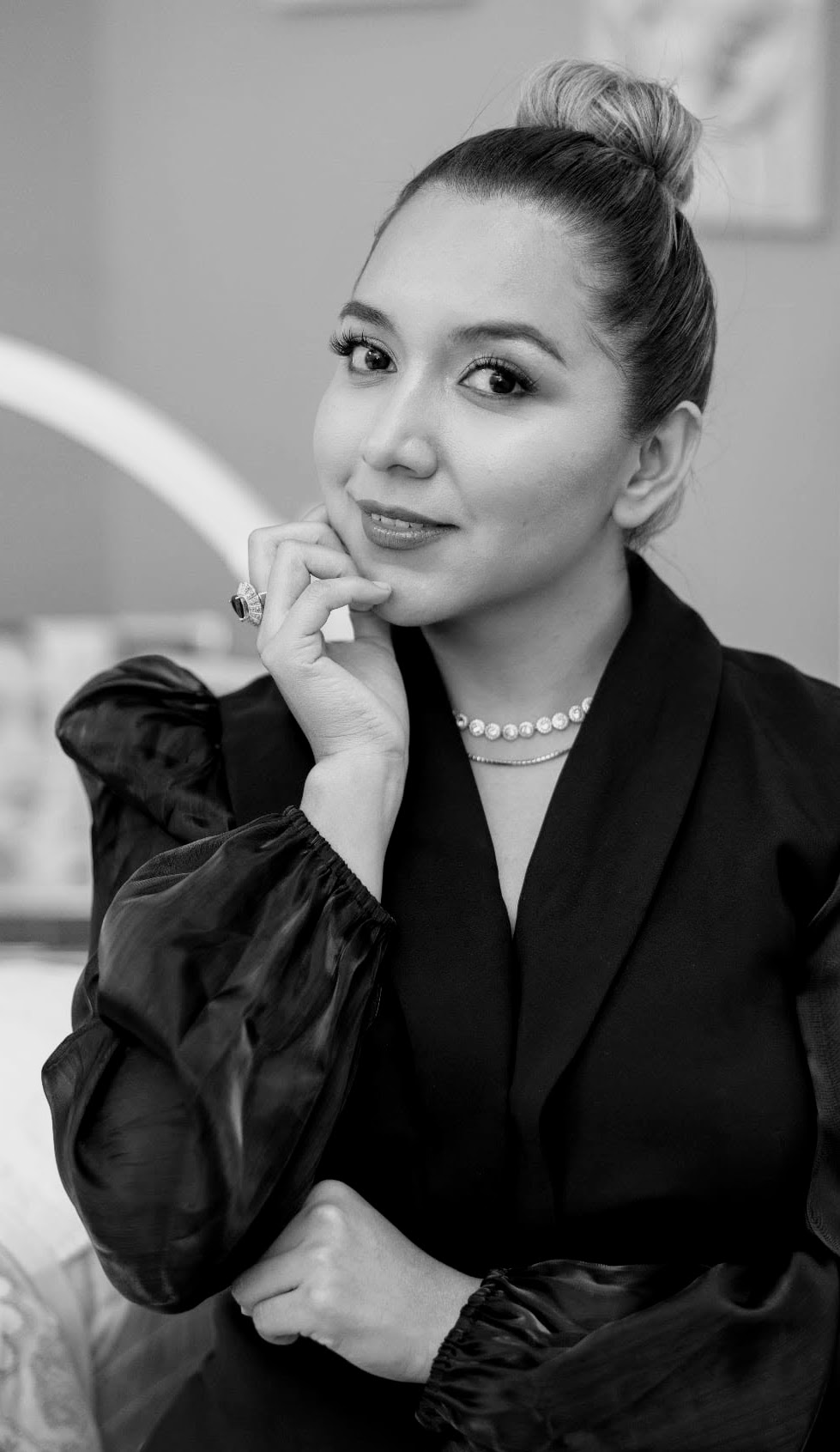 Woman with hair in a bun, wearing a dark blazer and pearl necklace, resting her chin on her hand.