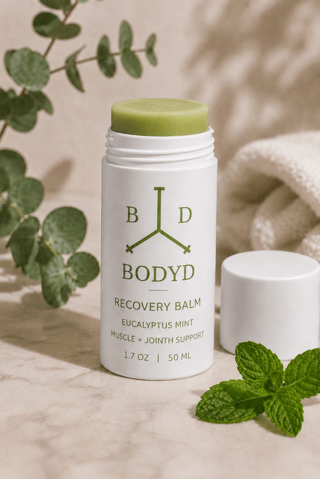 White tube of recovery balm with green lid, eucalyptus mint scent, on marble surface with green leaves nearby