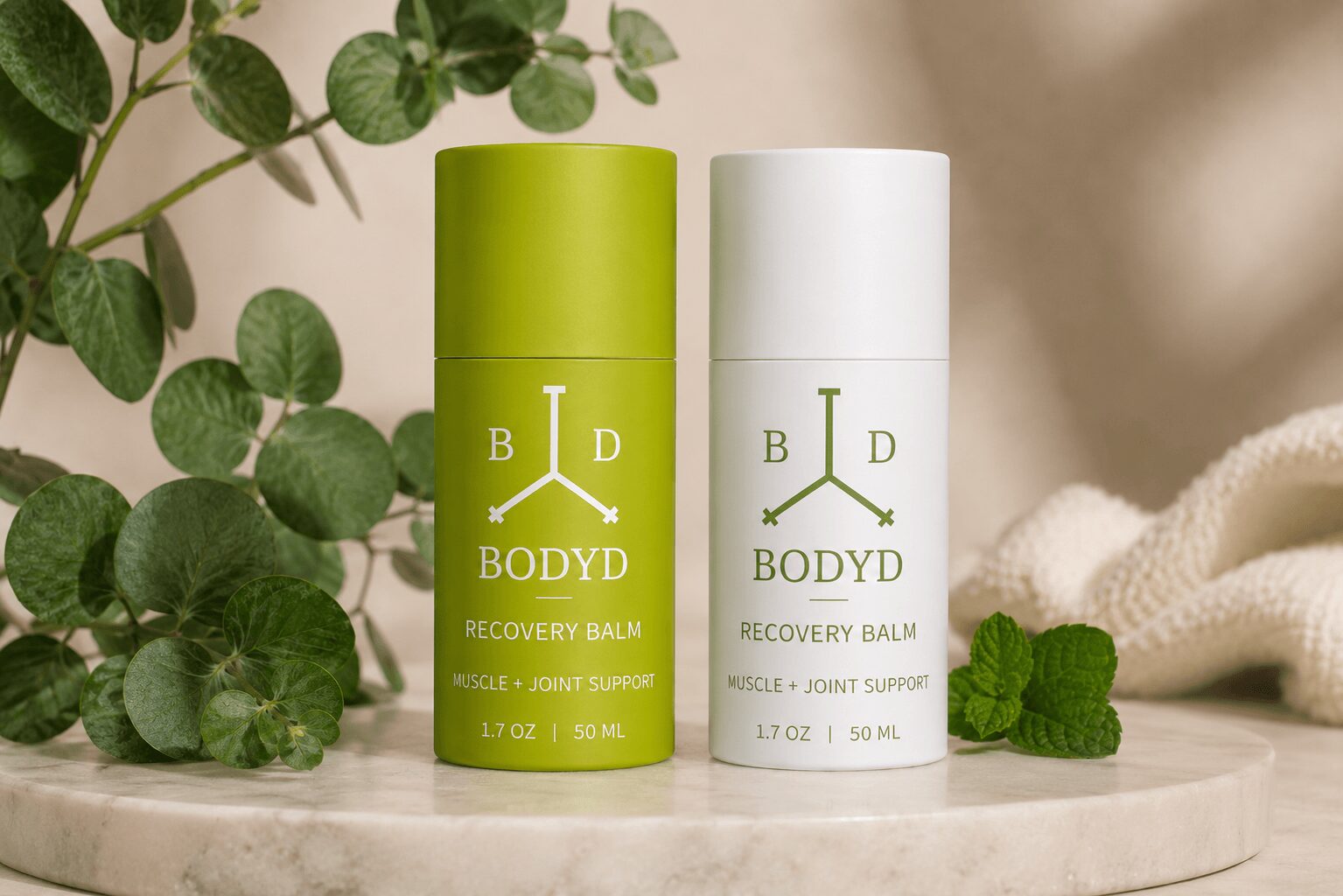 Two cylindrical containers of body balm on a white surface with green plants in the background, one green and one white.
