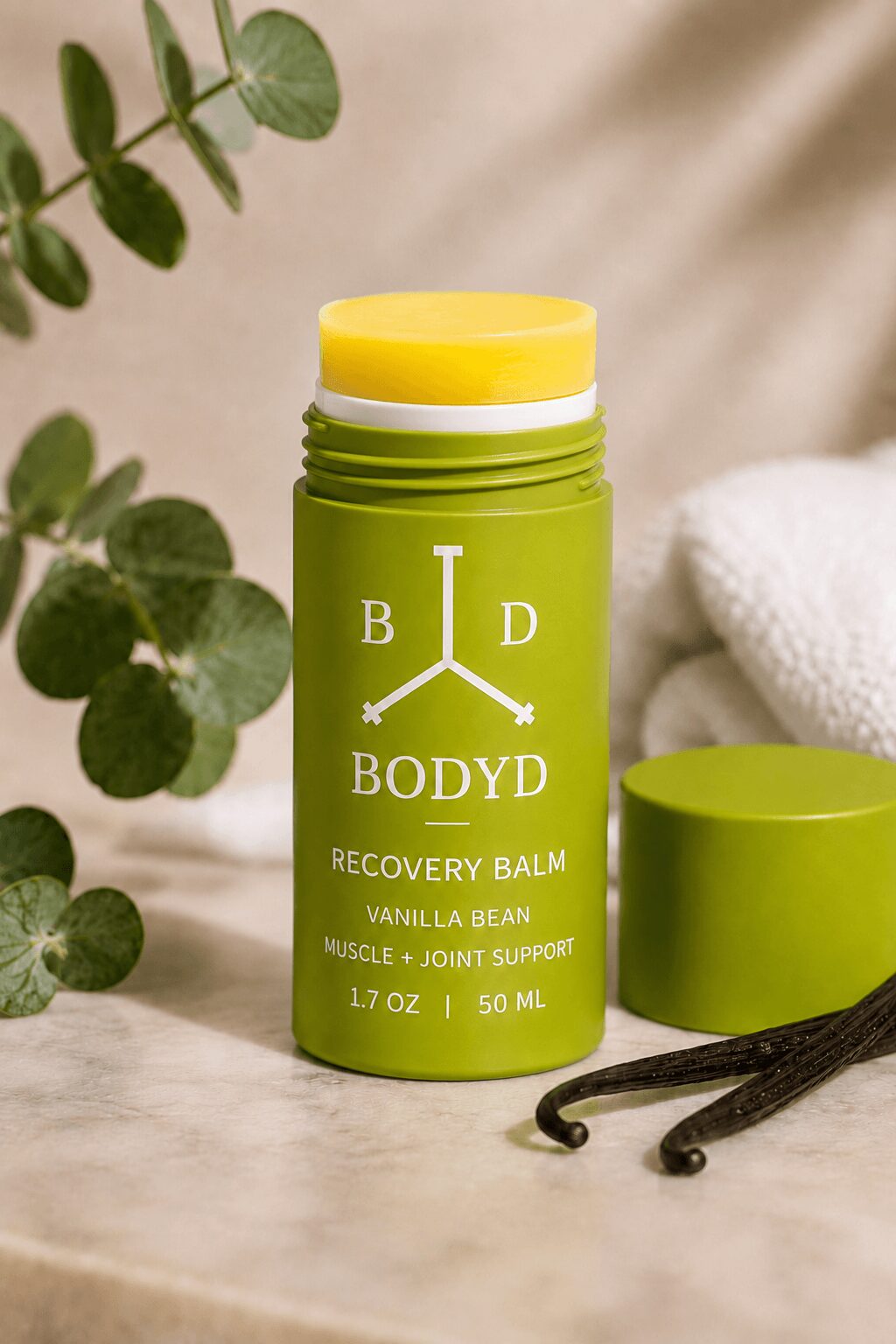 Green bottle of BodyD recovery balm with yellow lid, surrounded by eucalyptus leaves and vanilla beans, on a beige surface.