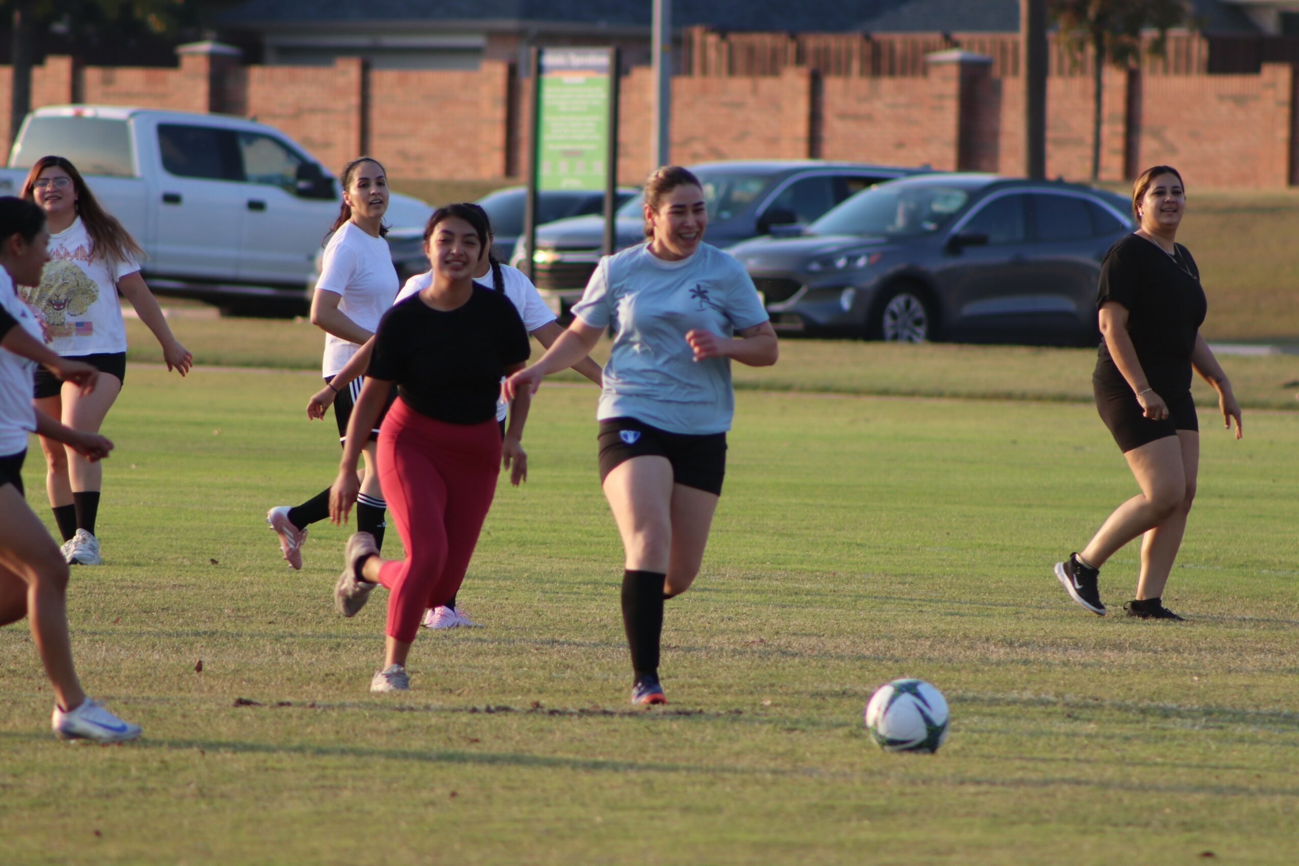 Group of women running on a grassy field with a soccer ball, cars parked in the background.