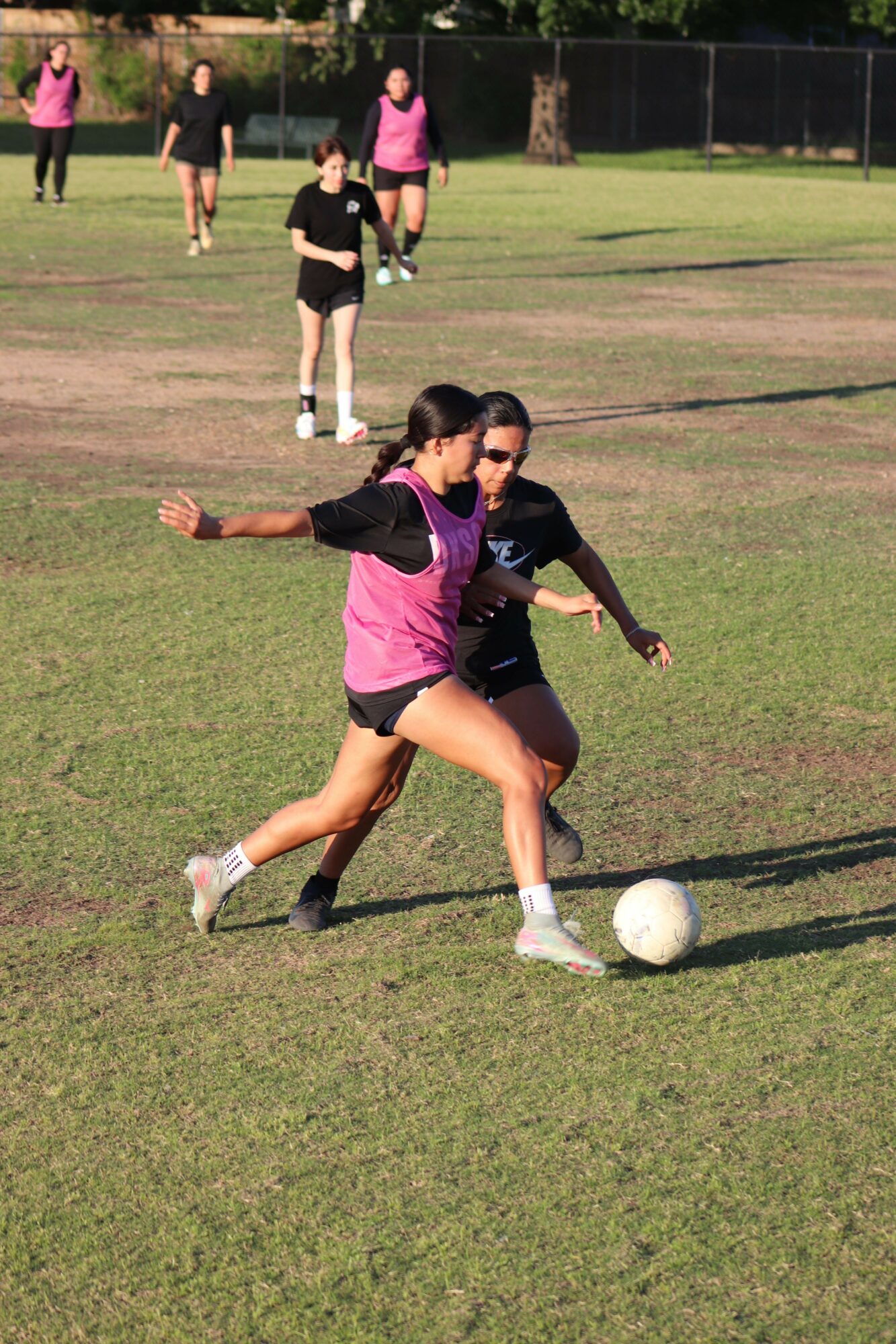 Person in pink and black sportswear kicking a soccer ball on a grassy field, with other people in the background.