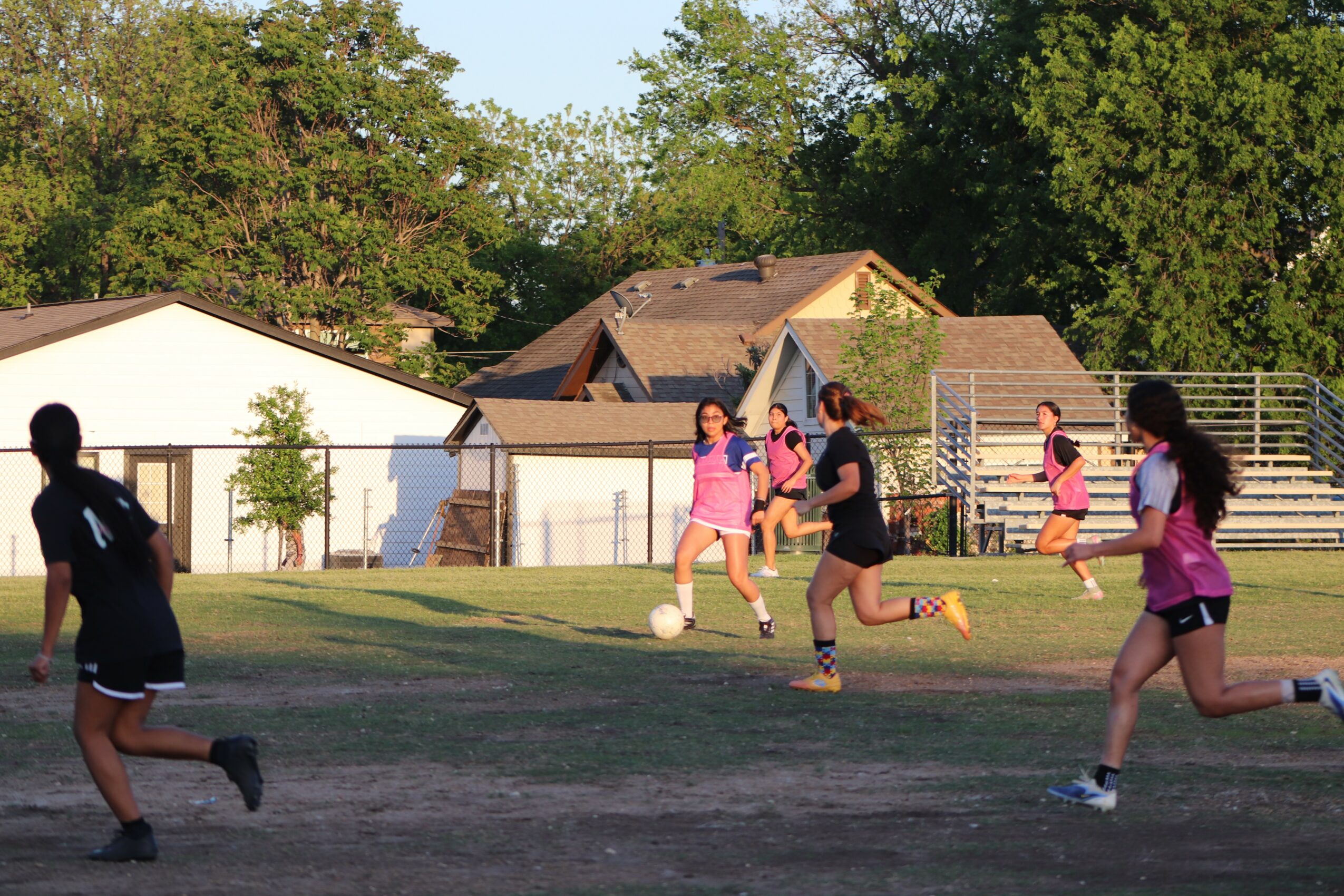 Group of children playing soccer on a grassy field with houses and trees in the background.
