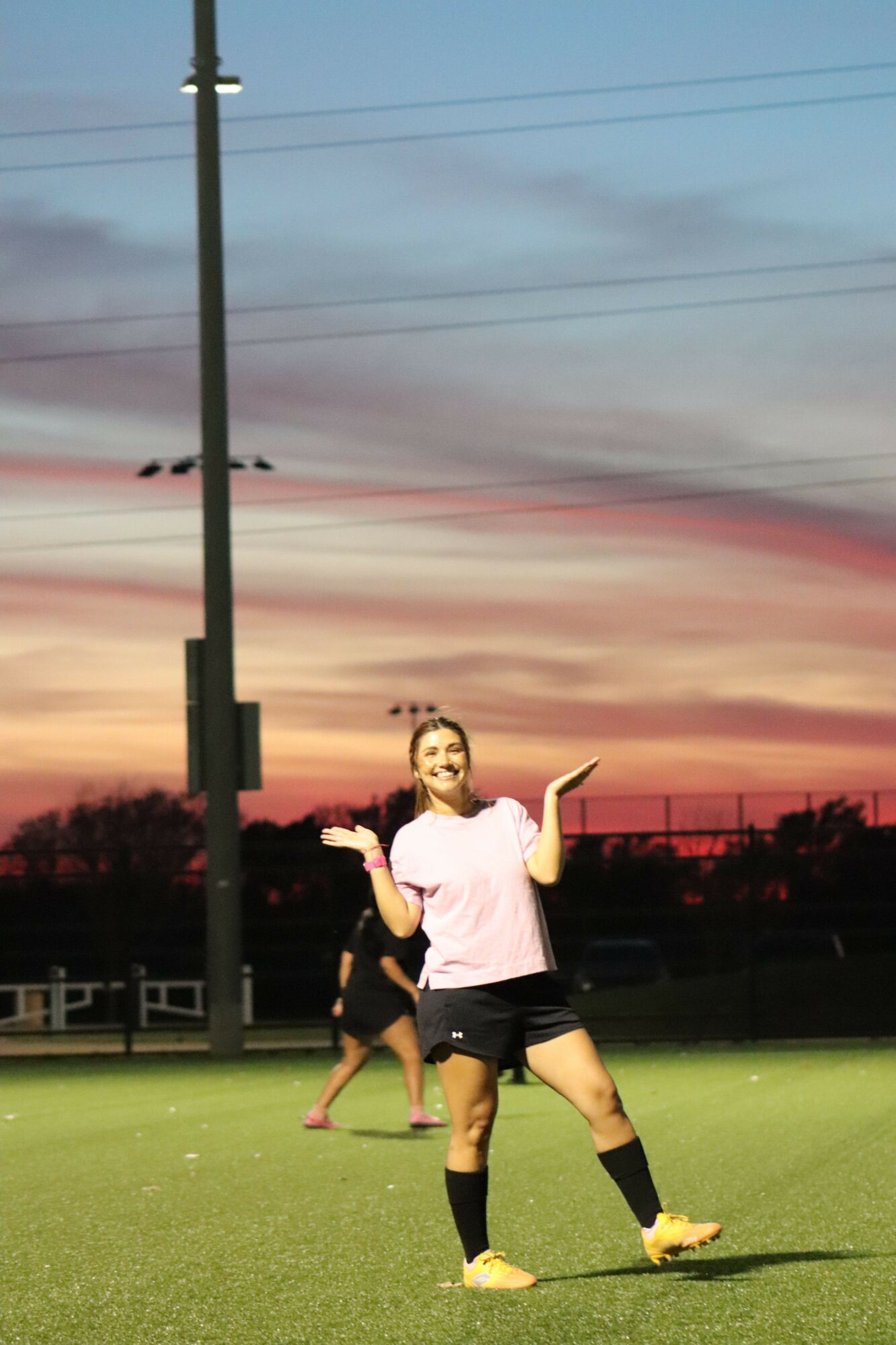 Person standing on grass field at sunset, wearing white shirt and black shorts, with a sunset sky in background.