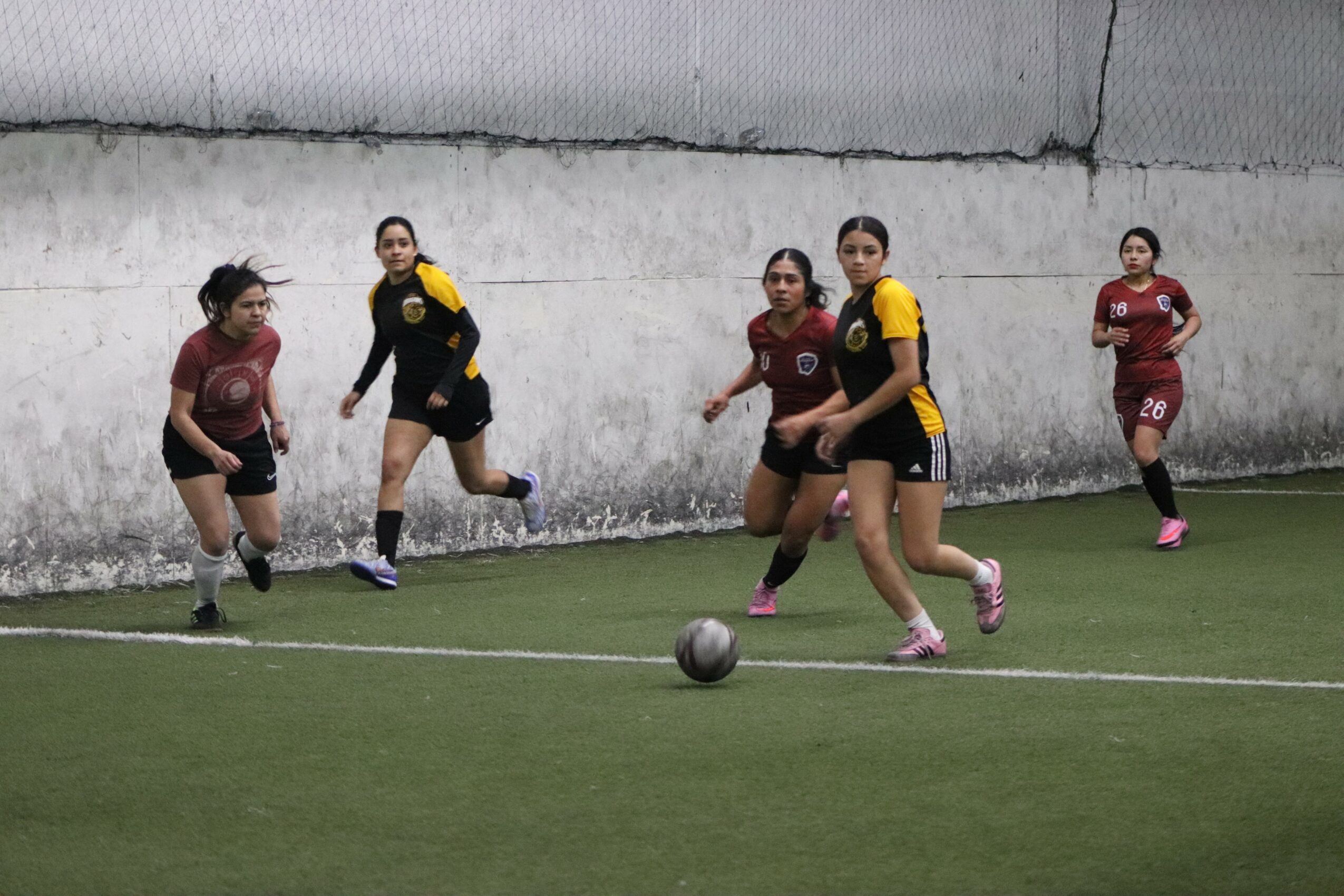 Four young women playing soccer on a field near a concrete wall, with one girl in yellow and black uniform controlling the ball.