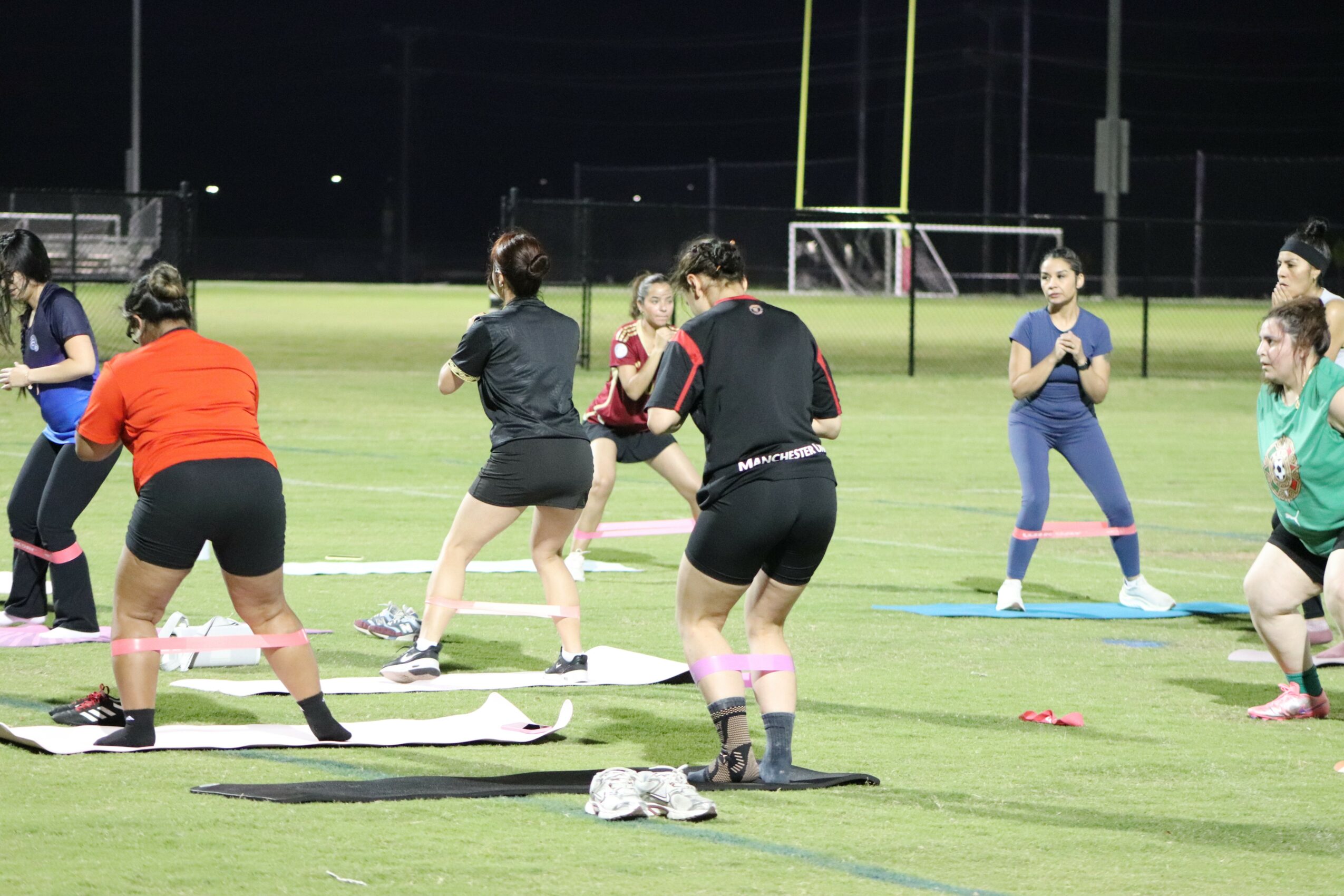 Group of people practicing exercises on a grassy field at night, some on mats, others standing or kneeling, with sports equipment nearby.