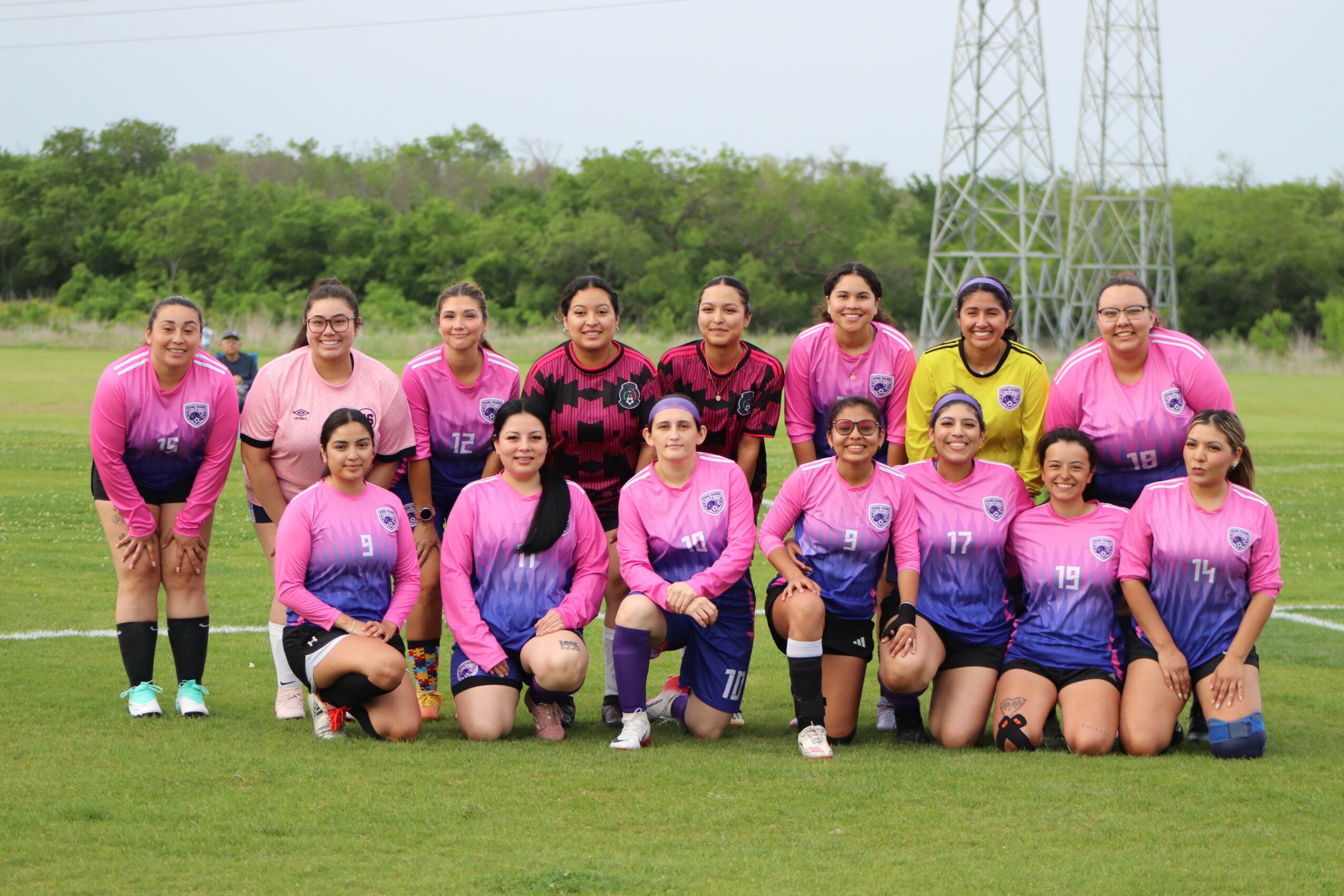 Group of 13 women in sports uniforms standing and kneeling on a grassy field, with trees and power lines in the background.