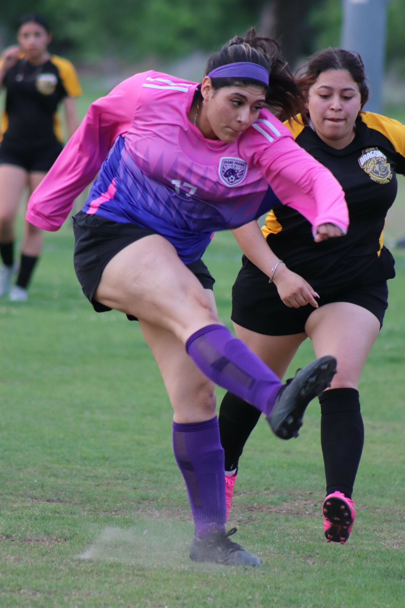 Two female soccer players in action on the field, one in pink and purple, the other in black and yellow, with a grassy background.