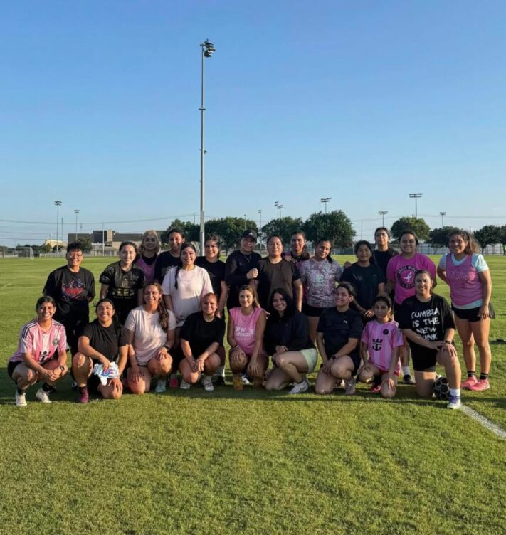 Group of people posing on a sports field with a clear blue sky, some standing and some kneeling, in casual clothing.