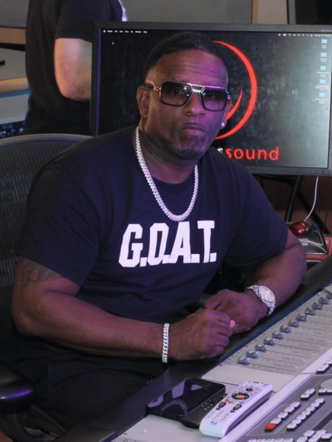 Man wearing sunglasses, a black GOAT T-shirt, and a pearl necklace, sitting at a sound mixing console in a studio.