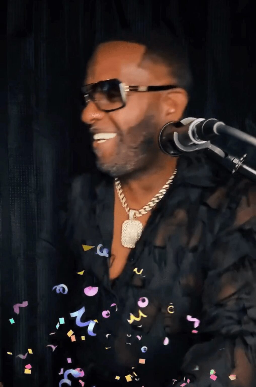 Man with sunglasses and a gold chain smiling near a microphone, dark background, colorful confetti around him.