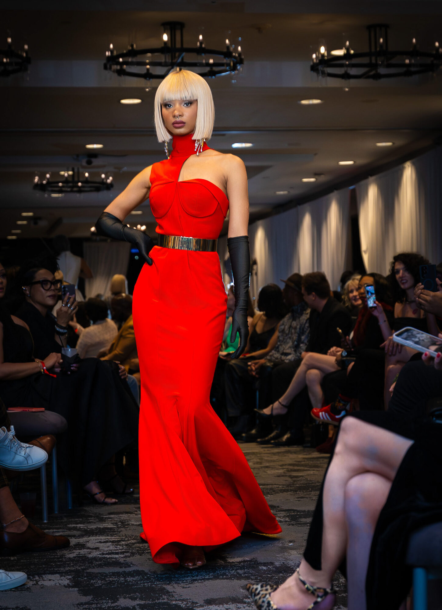 Woman in red gown with blonde bob hairstyle walking on runway at fashion show, audience seated on sides, dim lighting