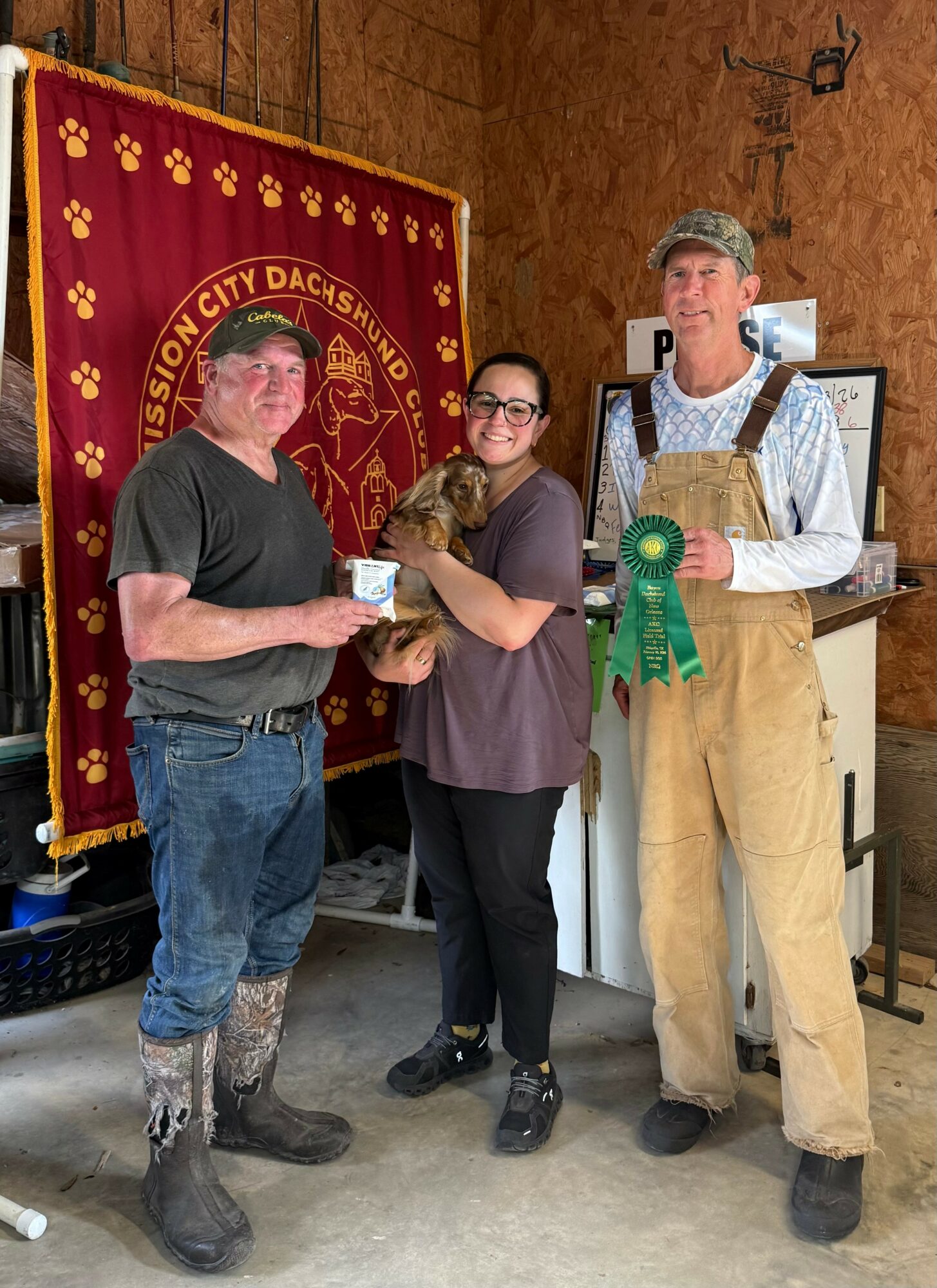 Three people standing indoors, one holding a small animal, with a red and gold banner in the background.