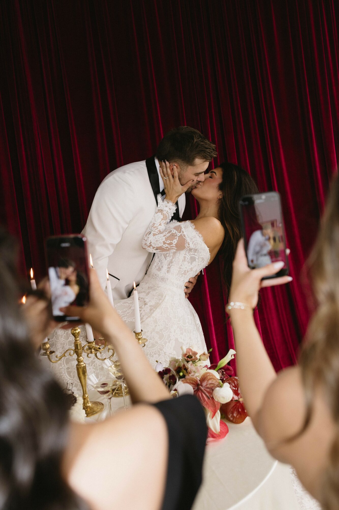 Couple kissing on stage with red curtain background, surrounded by people taking photos, wedding setting, woman in white dress.