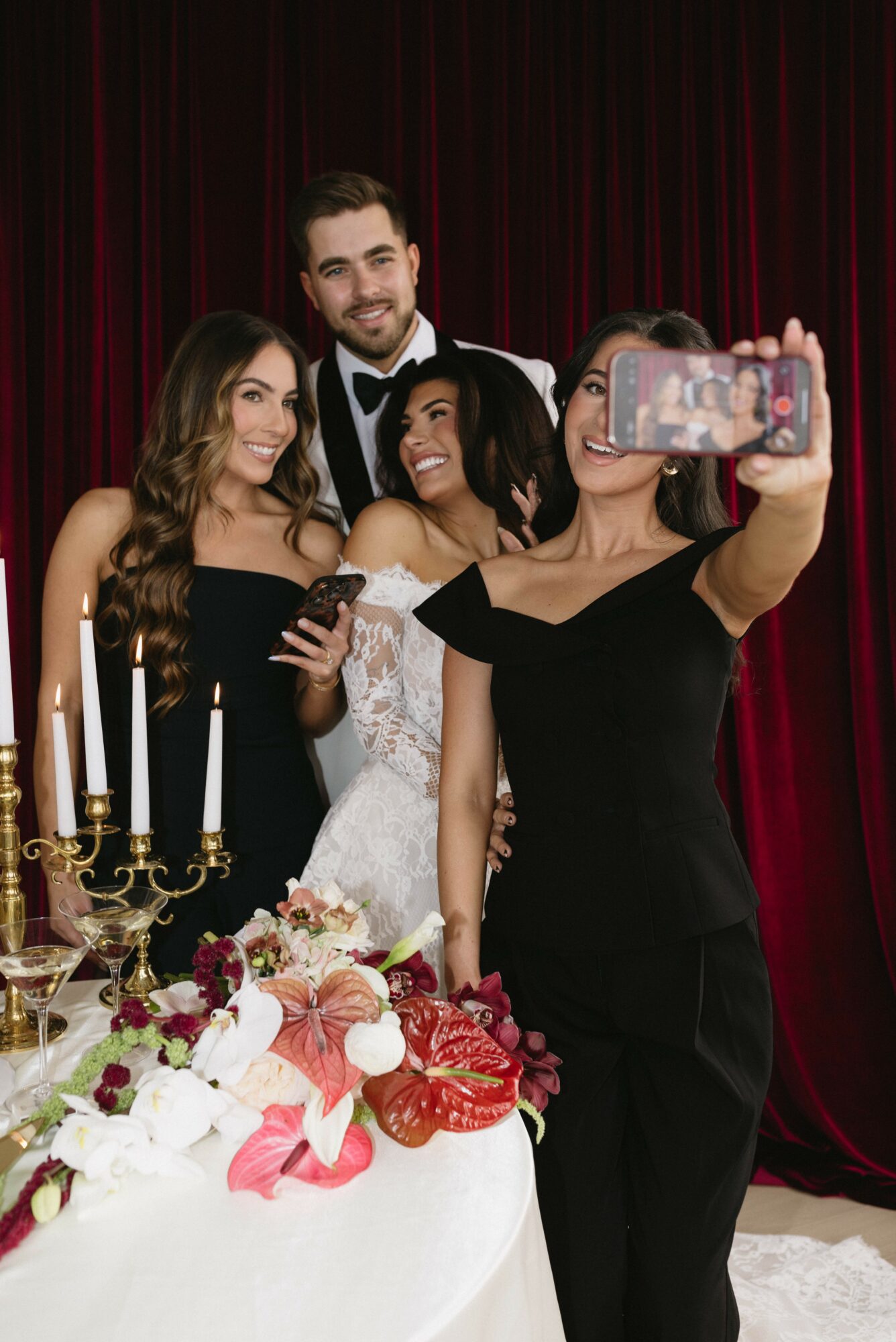 Four people, two women and two men, pose for a selfie at a celebration with a decorated table and red curtain background.