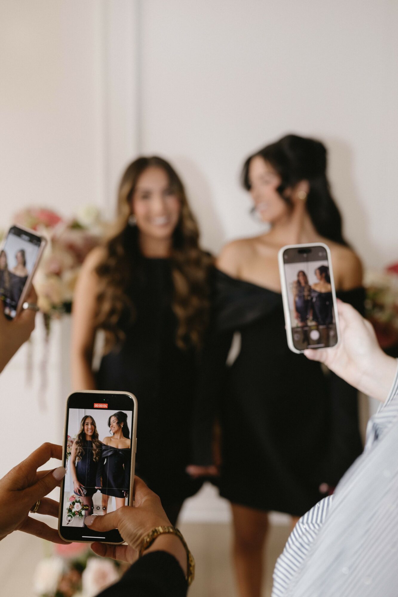 Two women in black dresses posing for photos, people taking pictures with smartphones, indoor setting, smiling faces.