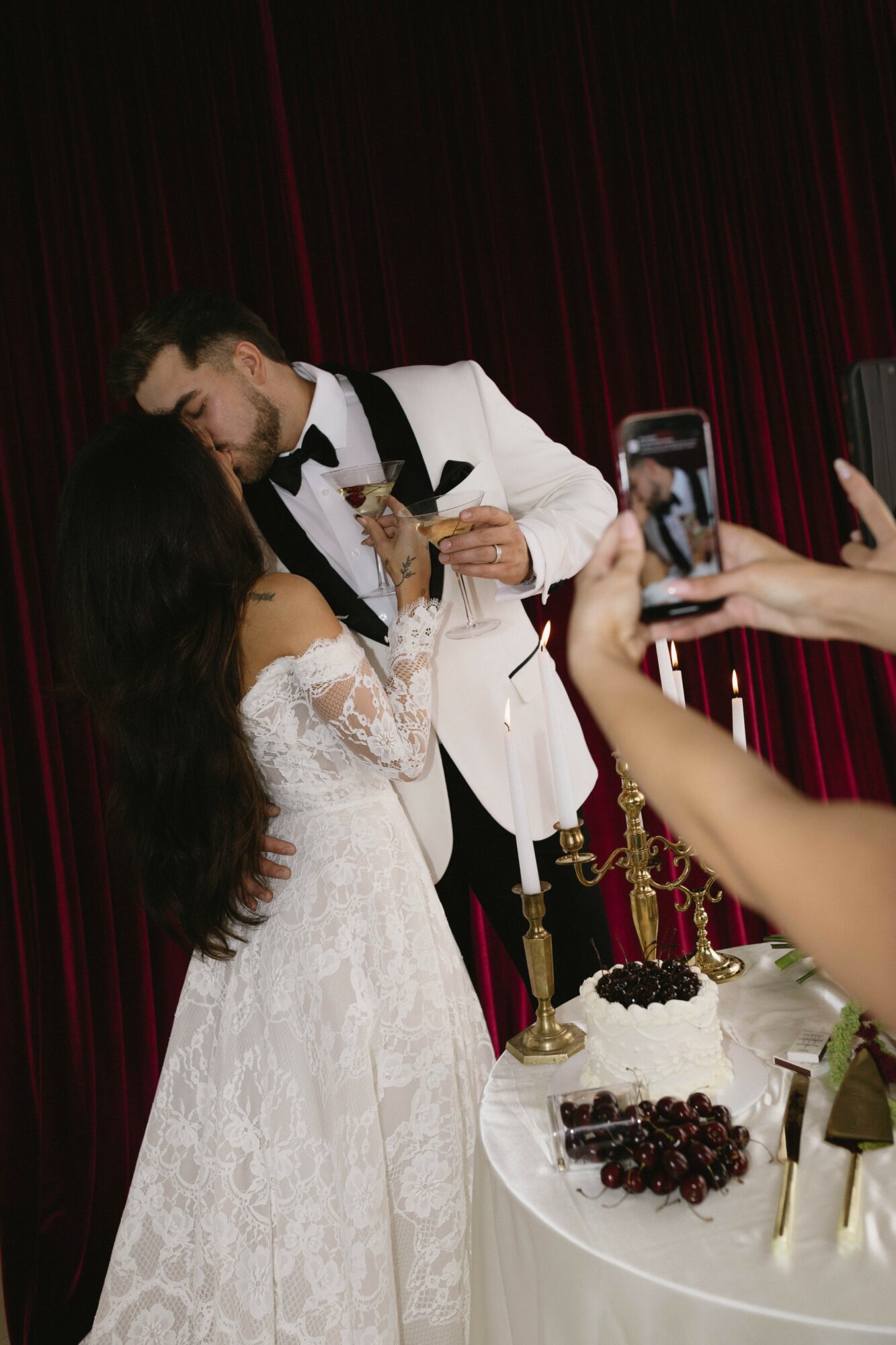 Couple in wedding attire sharing a moment, with a person taking a photo, and a table with grapes and candles in the foreground.