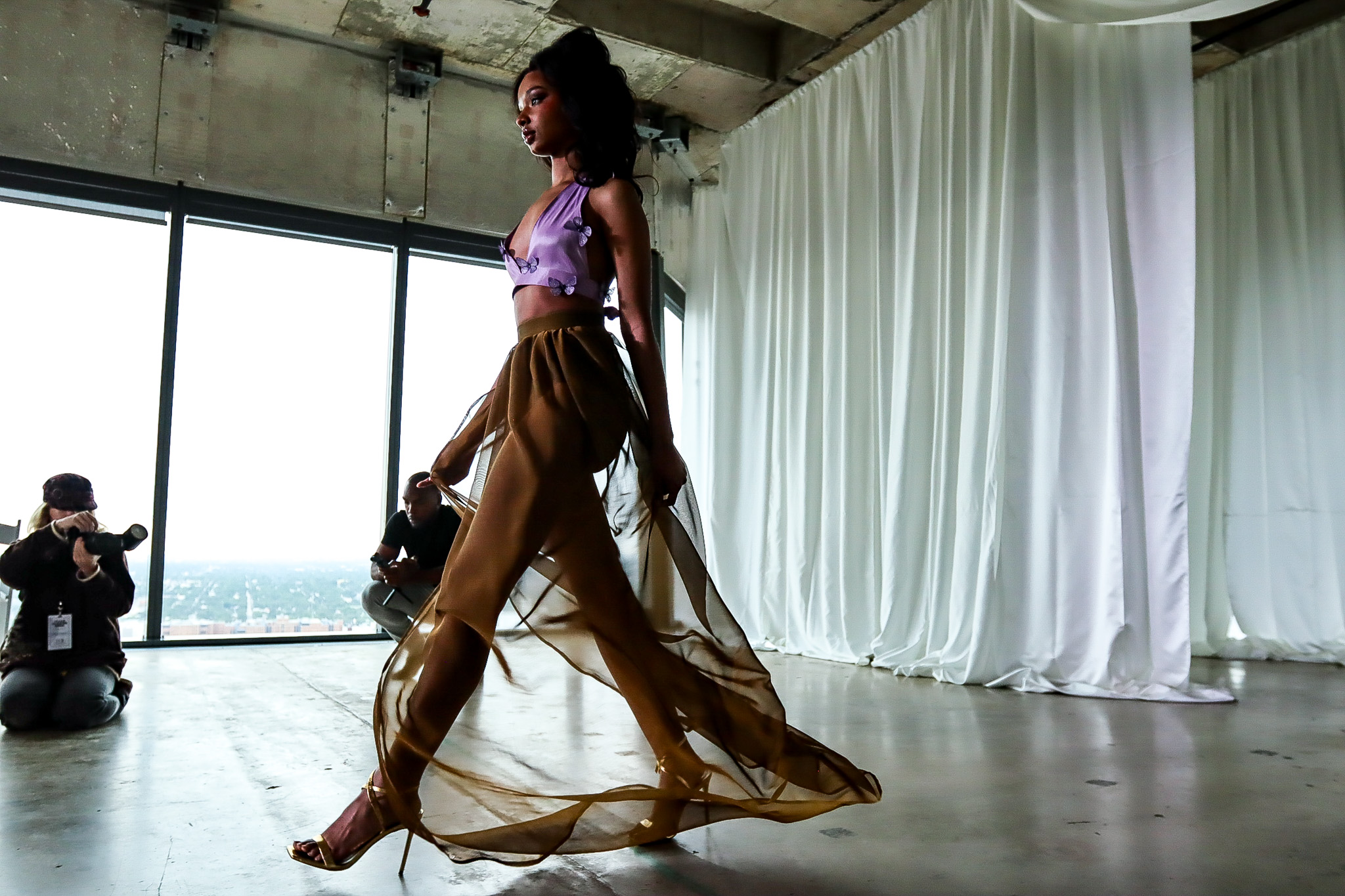 A woman walks in a spacious room with large windows and white curtains, with photographers capturing her from the side.